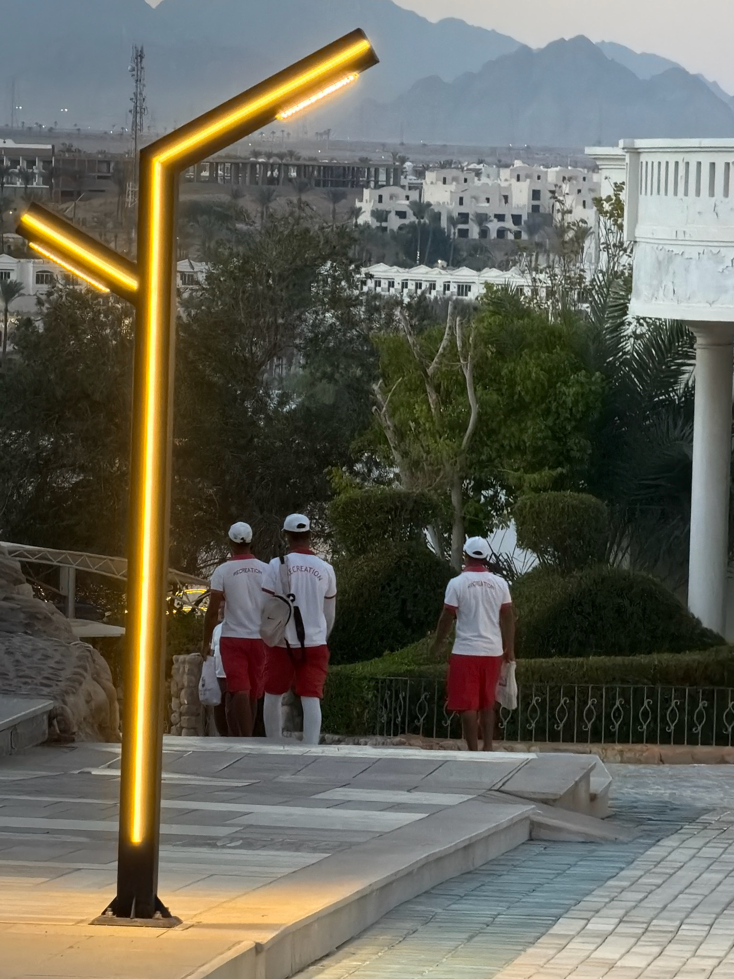 This image depicts a scene in Sharm El Sheikh, Egypt, likely at a resort or hotel complex. In the foreground, a modern, angular lamppost with integrated yellow LED lighting stands prominently. A paved pathway with a textured surface leads upwards, bordered by stone landscaping on the left and manicured hedges and trees on the right. In the middle ground, three individuals, presumably staff members, are walking away from the camera up the pathway. They are all wearing white caps, white t-shirts with the word "RECREATION" printed on the back in red lettering, and red shorts. Two of them also carry white bags. Behind them, a white building with a colonnaded structure is partially visible on the right, and further in the background, a cluster of white buildings with flat roofs is set against a backdrop of hazy mountains. The overall atmosphere suggests an evening or early morning setting, given the artificial lighting and the subdued natural light.