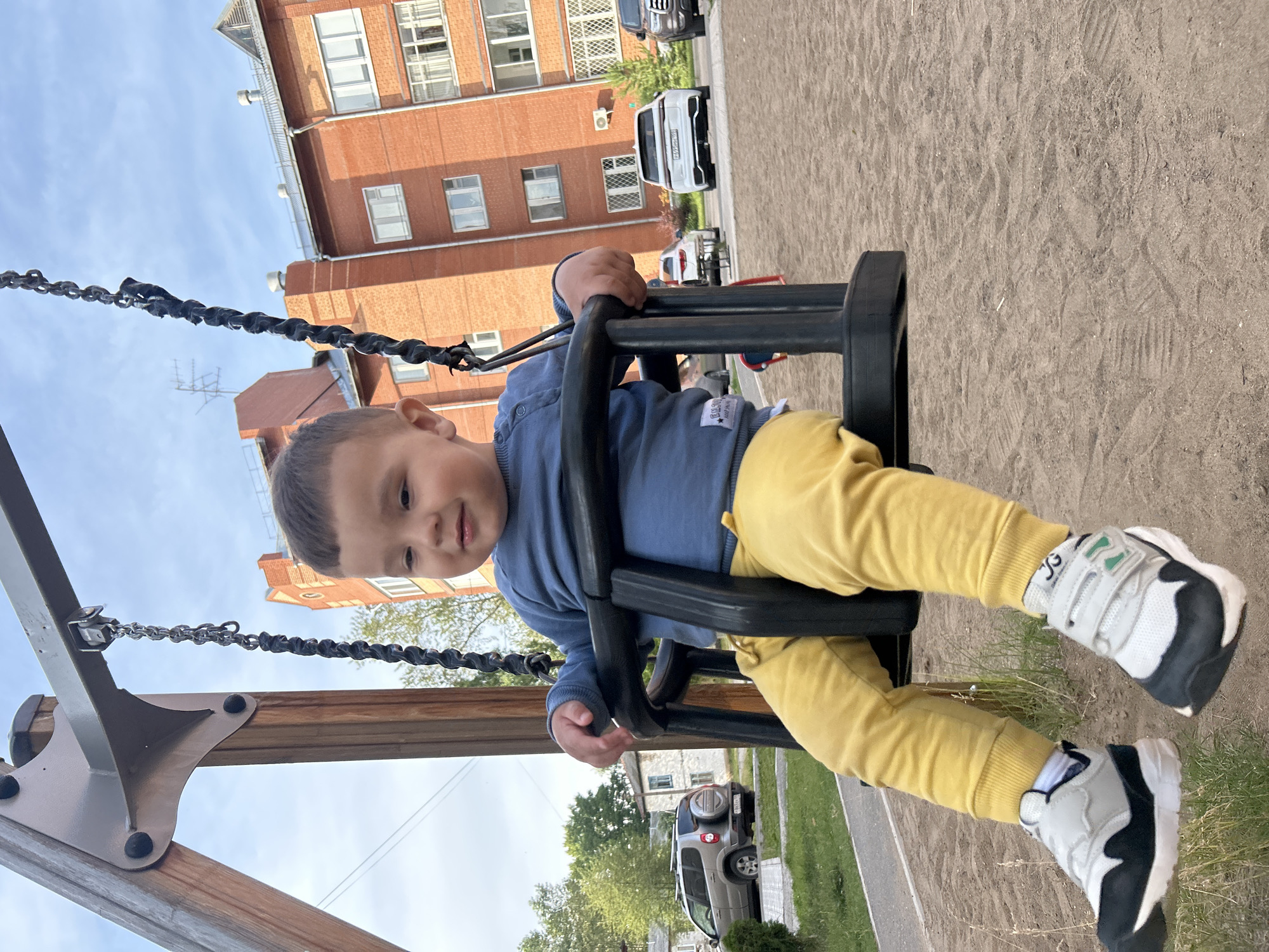 A young boy with dark hair and brown eyes is sitting on a swing set in a playground. He is wearing a blue long-sleeved shirt and yellow pants. His feet are clad in grey and black sneakers with white laces. The swing set has black chains and a black seat.
In the background, there is a tall brick building with many windows and a white-tiled roof. Several cars are parked in front of the building, including a white SUV and a silver SUV. The ground of the playground is covered in sand, with some grass and trees visible beyond the sand. The sky is a light blue with some clouds. The image appears to have been taken during the daytime.
The boy is looking towards the camera with a slight smile, suggesting he is happy and enjoying himself. The overall scene depicts a typical day at a playground, with a child playing on a swing. The building in the background suggests an urban or suburban setting. The time of day appears to be late morning or early afternoon, given the angle of the sun and the brightness of the sky. The weather appears to be pleasant.