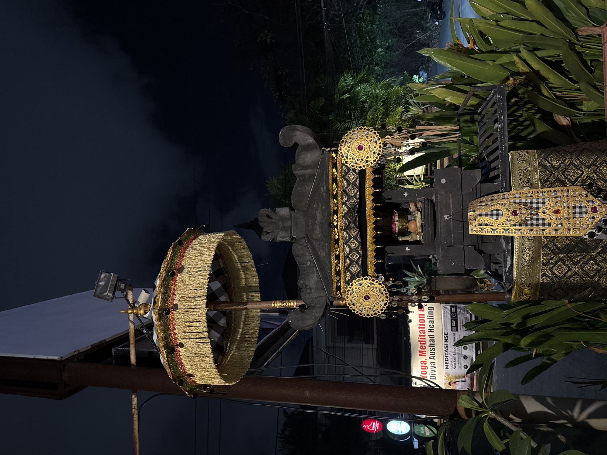 This is a night-time photograph, likely taken in Kuta, Indonesia, featuring a decorative structure with ornate gold and black fabric elements. A large, circular, golden umbrella-like object with fringed edges is prominently displayed on the left side of the image, supported by a dark pole. Behind it, a dark, carved stone structure is visible, adorned with gold decorative elements and red gemstones.
To the right of the stone structure, more of the gold and black fabric is visible, suggesting it's part of a larger decorative display. A white sign with text in various colors is partially visible in the lower right quadrant. The visible text includes "Yoga, Meditation & Divya Aushad Healing" and "MEDITASI NSE Neo Self-Empowerment".
Lush green foliage surrounds the decorative elements, and the background is dark, indicating it's nighttime. A faint light source from a building or streetlamp is visible in the upper left corner, illuminating a portion of a roof. The overall atmosphere is festive and suggests a cultural or spiritual setting, possibly a temple entrance or a special event. The lighting conditions and the presence of decorative elements point to an evening or night-time scene.