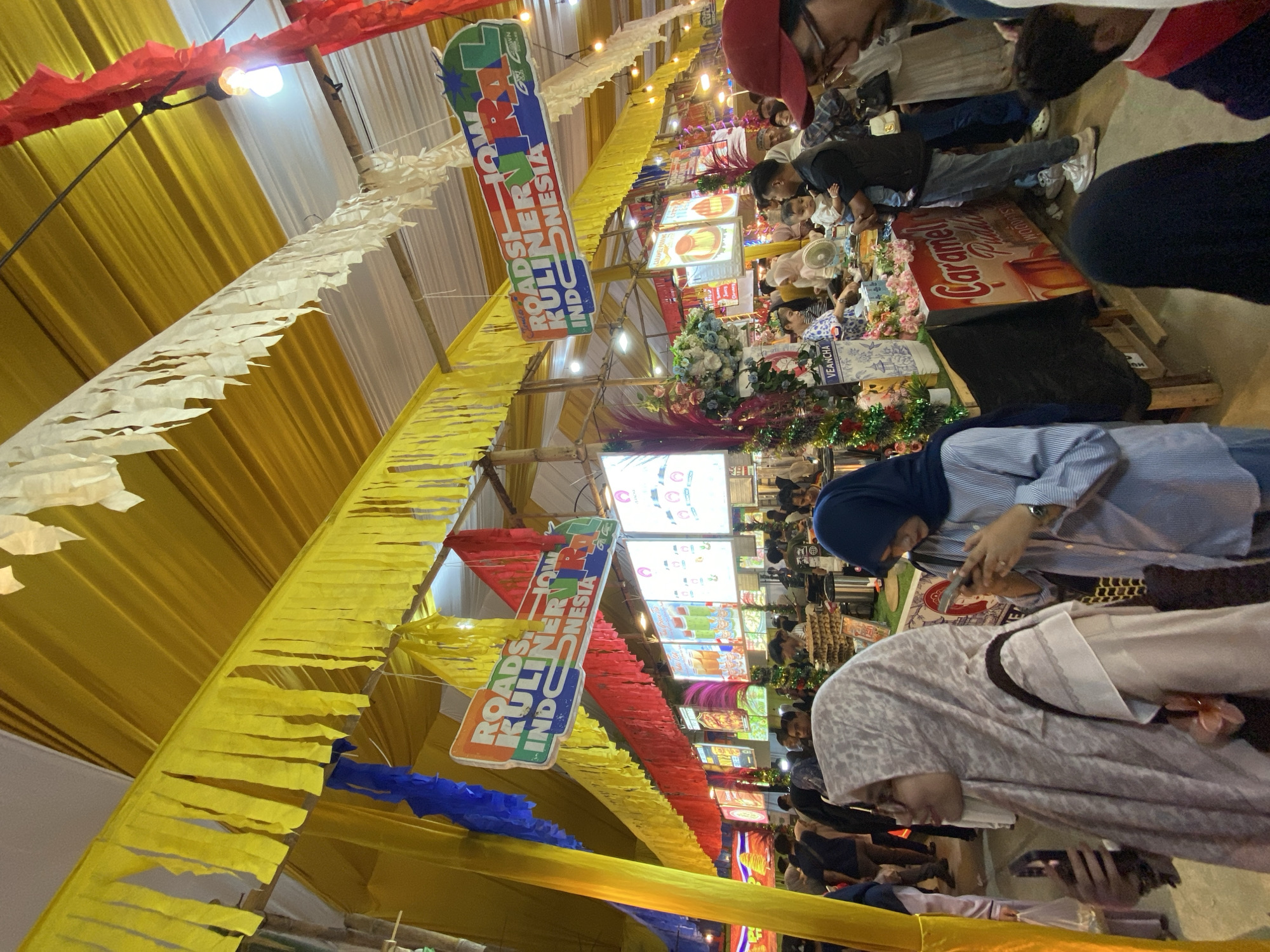 This media file captures a vibrant scene at a food festival, likely in Lhokseumawe, Indonesia. The focus is on a lively market atmosphere with numerous stalls and attendees. The overall impression is one of a busy and engaging event.
The scene is decorated with colorful streamers, primarily in yellow, red, and white, creating a festive ambiance. Several signs are visible, including one that reads "ROADSHOW KULINER INDONESIA," indicating a culinary event showcasing Indonesian food. Another sign highlights "Caramel" and "Puding Karamel," suggesting a dessert offering.
Several people are visible, engrossed in various activities. Some are browsing the stalls, while others appear to be interacting with vendors or friends. The individuals are dressed in casual attire, and some women are wearing hijabs. The presence of children suggests a family-friendly environment.
The lighting suggests it might be daytime or early evening, as there is a mix of natural and artificial light from the overhead lamps. The overall mood appears to be cheerful and energetic, typical of a public festival. The layout suggests an indoor or semi-outdoor venue, possibly a large hall or an open-air market area covered with tents. The presence of food stalls, crowds, and festive decorations strongly points to a food-related event.