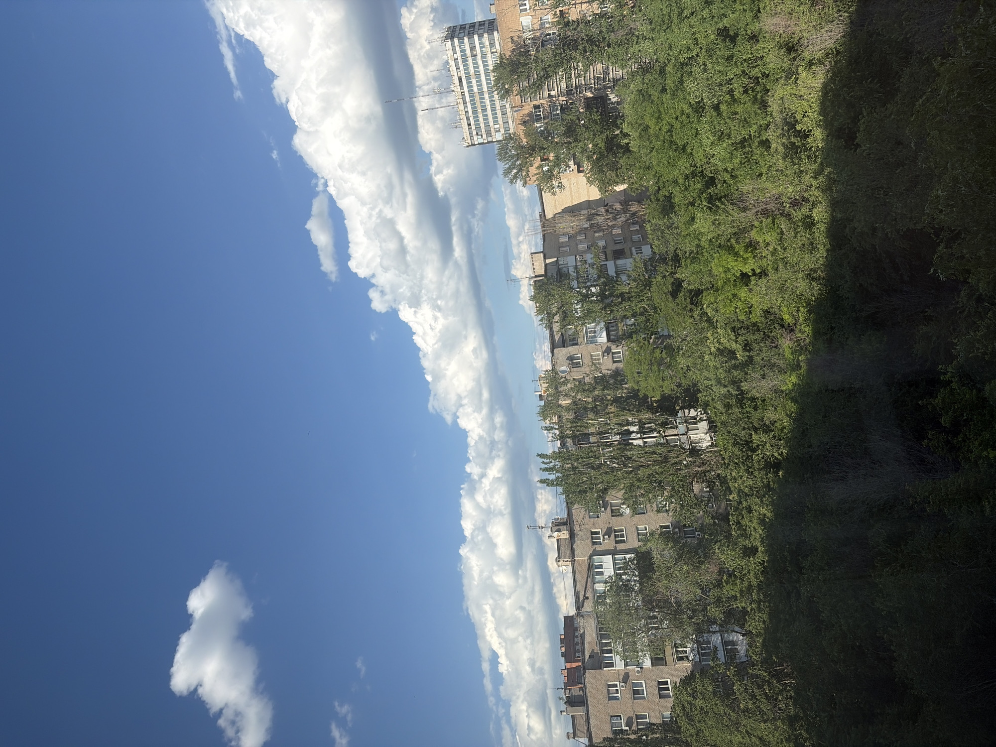 The image displays a cityscape with a prominent blue sky filled with fluffy white clouds. A row of apartment buildings, interspersed with lush green trees, runs along the right side of the frame. The buildings appear to be of varying heights and architectural styles, with visible windows and balconies. Some of the trees are tall and dense, casting shadows on the buildings and the foreground. The sky is bright and clear, suggesting it is daytime, likely a sunny afternoon given the shadows. The clouds are dramatic and voluminous, creating a sense of depth and movement in the sky. The scene is a typical urban environment, with a blend of man-made structures and natural elements. There is no discernible activity or event taking place, and no visible text in the image. The overall impression is one of a calm and pleasant day in a city.