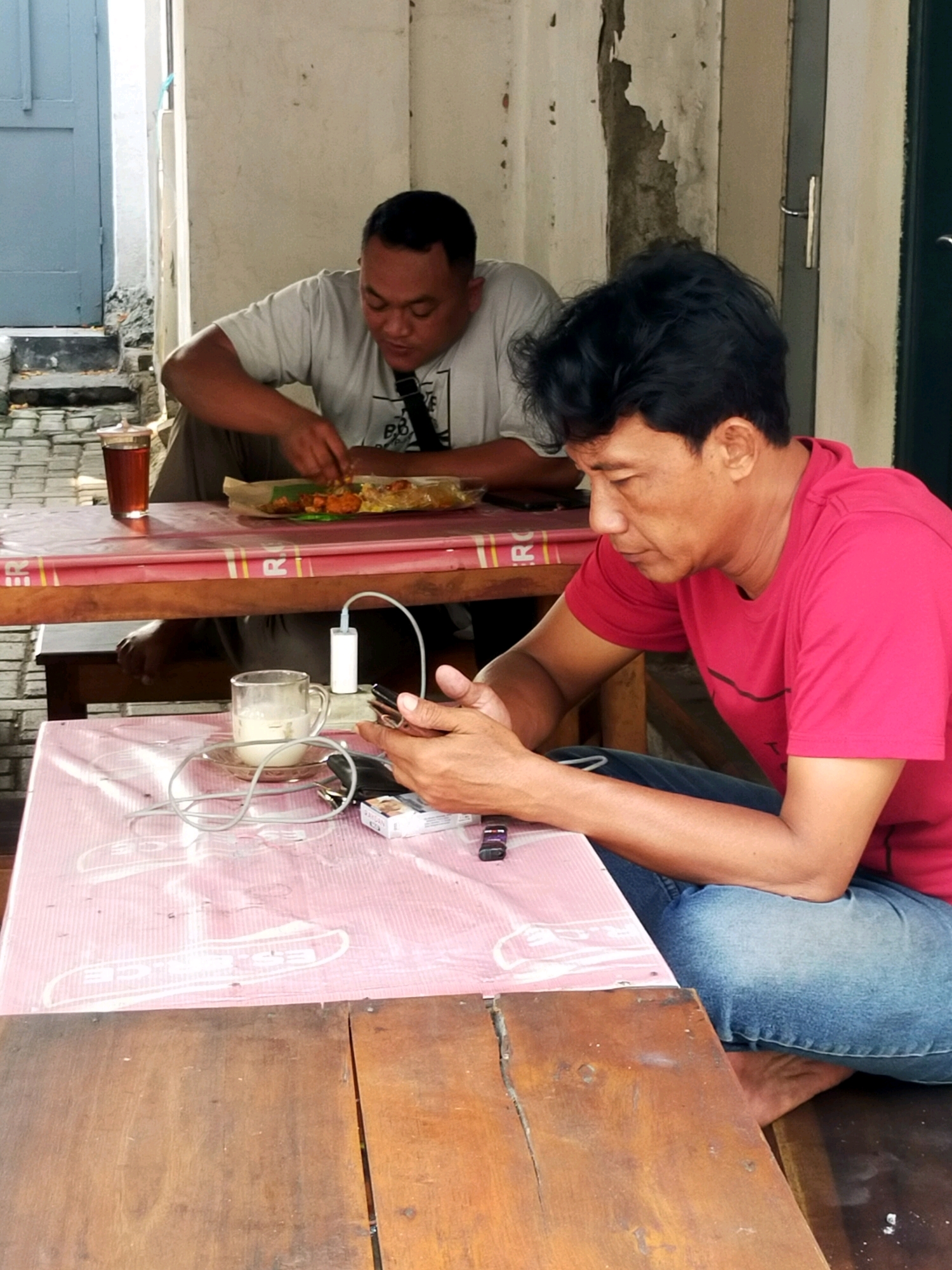 The image depicts two men sitting at separate tables outdoors. The man in the foreground, wearing a pink t-shirt and blue jeans, is looking down at his mobile phone and holding it in his hands, which are resting on a pink, plastic-covered table. A charging cable connects to his phone and runs to a white power adapter. A glass of milk, a cigarette pack, and a lighter are also on the table in front of him. The man in the background, wearing a grey t-shirt and brown pants, is seated at a wooden table, eating from a plate of food wrapped in paper. He is looking down at his food and using his hands to eat. Behind the man in the background, a glass of iced tea sits on the table. The environment appears to be an open-air eating area, possibly a warung or roadside cafe, with concrete walls and a weathered facade. The setting suggests a casual dining or resting spot. The image appears to be taken in Surabaya, Indonesia, given the cultural context and common use of similar food packaging.