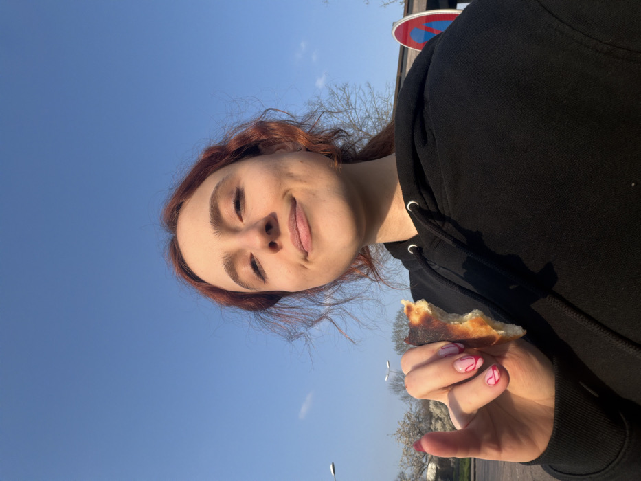 A young woman with reddish-brown, wavy hair, wearing a black hooded sweatshirt, is depicted in an outdoor setting in the city of Toul, France. She is seen from the chest up, looking directly at the camera with a faint smile. Her left hand, adorned with pink fingernails featuring white swirl patterns, holds a partially eaten piece of food, resembling a bread crust or pizza edge. The background features a clear blue sky with minimal clouds. To the left, two grey streetlights are visible, along with bare trees and some white blossoms. To the right, a circular red and blue road sign, consistent with a "no stopping" or "no parking" symbol, is partially observed behind the woman. The ground visible in the lower left background appears to be a paved surface.