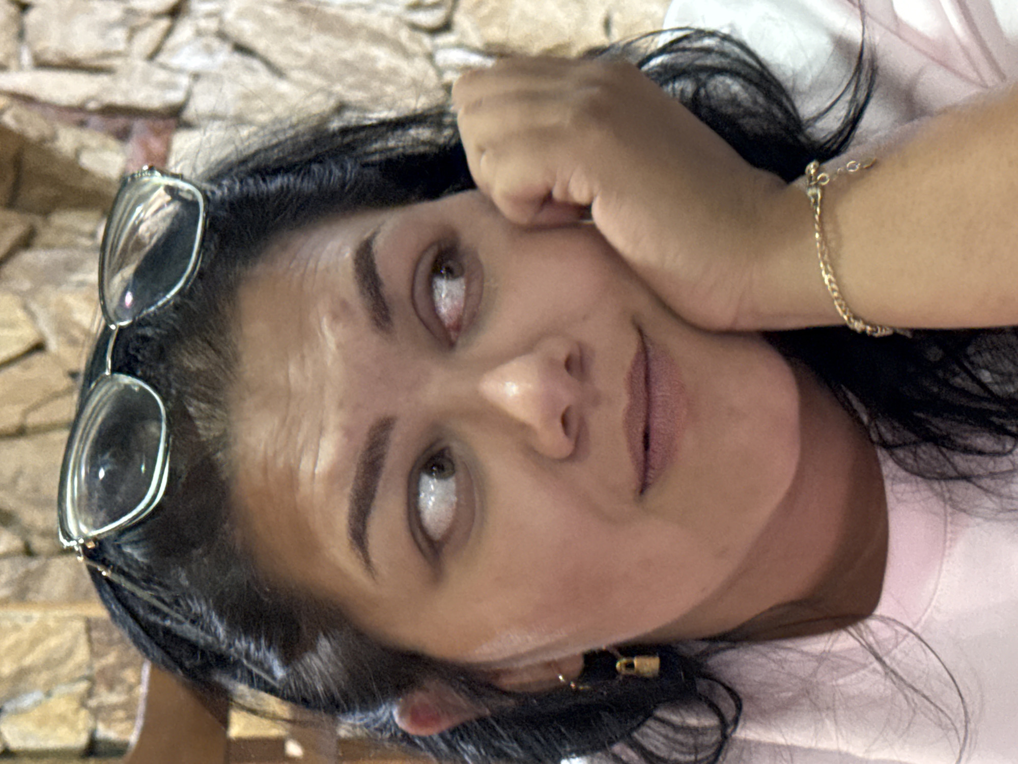 A medium close-up shot shows a woman from the chest up, looking to her left. She has dark hair pulled back, and she is wearing glasses perched on top of her head. Her right hand is resting on her cheek, and she has a gold bracelet on her left wrist. Her head is tilted slightly to her left. The background is a textured wall. The image was taken in Sredets, Bulgaria.