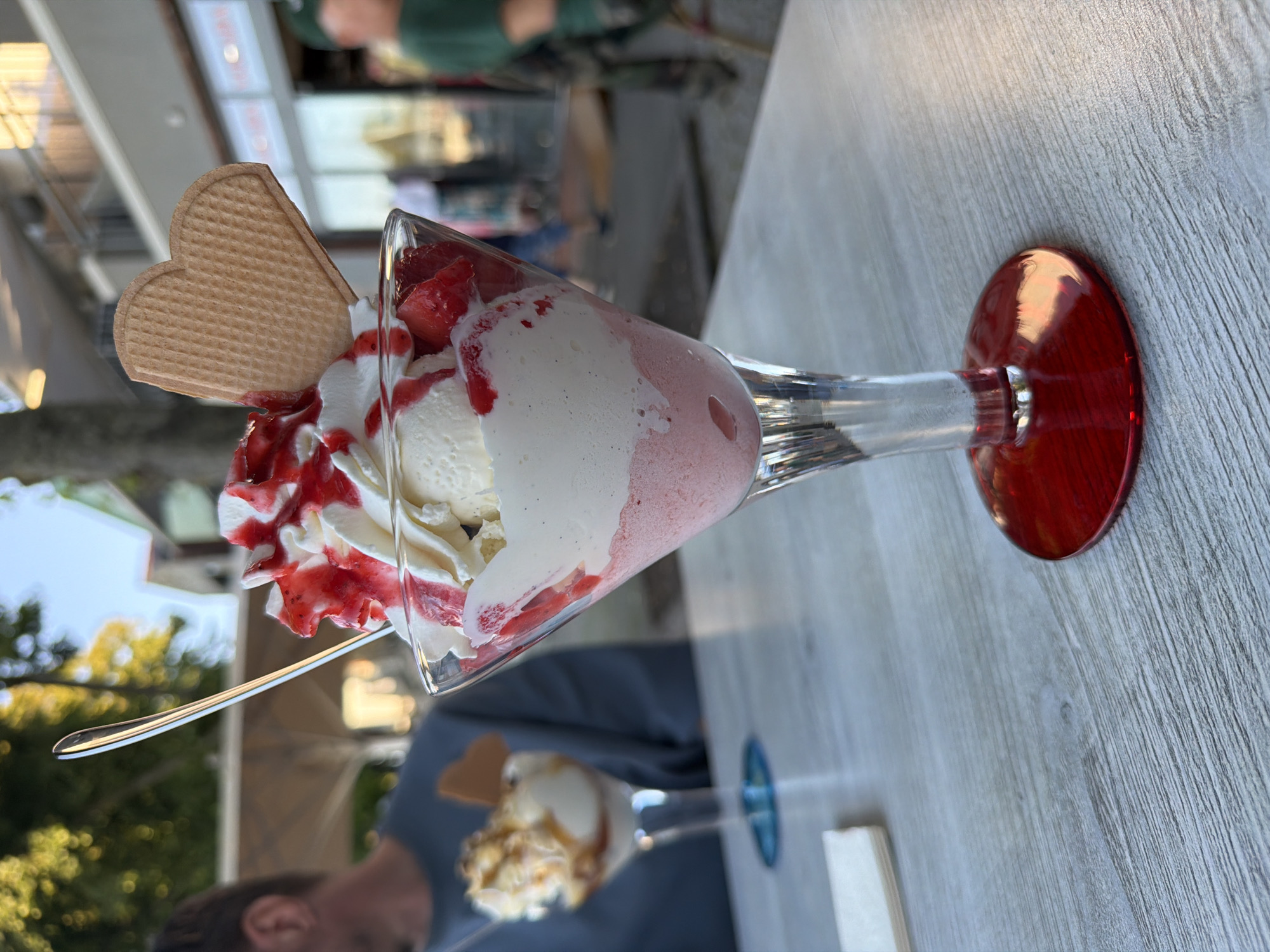 A strawberry sundae is presented in a stemmed glass on a light-colored wooden table. The sundae consists of layers of pink strawberry ice cream and white vanilla ice cream, topped with whipped cream and red strawberry syrup. A heart-shaped waffle cookie is inserted into the whipped cream. In the background, a person wearing a blue shirt is visible to the left, and another person in a green shirt is to the right, partially out of focus. The setting appears to be an outdoor cafe in Rostock, Germany, with trees and buildings visible in the distance under a clear sky.