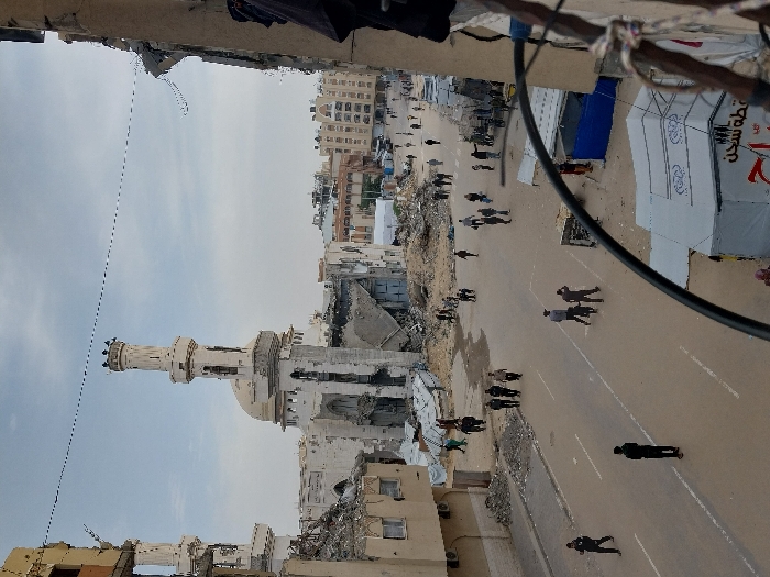 The image depicts a wide-angle view of a heavily damaged urban street in Khan Yunis, Palestinian Territory, under an overcast sky. In the background, a prominent mosque with a central dome and two minarets stands significantly damaged, with large sections of its structure missing or collapsed. Surrounding the mosque and extending along the street are numerous other buildings, many showing extensive structural damage, exposed interiors, and piles of rubble at their bases.
The paved street, marked with white lines, is populated by approximately two dozen individuals, mostly men, walking or standing in various small groups. On the right side of the street, closer to the foreground, are makeshift structures including a large white tent with Arabic writing and a smaller structure covered by a blue tarp, indicating temporary shelters or market stalls amidst the destruction. Piles of debris and rubble are visible along the edges of the road and within damaged building footprints. A thick, black curved cable hangs into the lower right foreground, partially obstructing the view, suggesting the photograph was taken from an elevated position within a damaged structure. Another overhead utility line spans the upper portion of the frame.