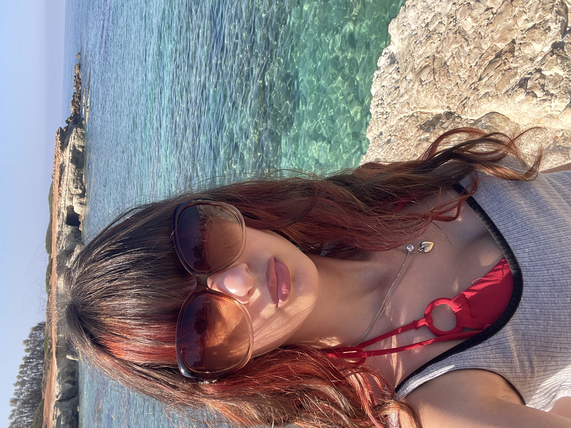 In Peyia, Cyprus, a young woman with auburn-streaked brown hair and large sunglasses poses for a selfie near the sea. She is wearing a red bikini top under a gray tank top and a necklace with a heart pendant. The background shows the clear blue Mediterranean Sea, rocky cliffs, and a clear sky.