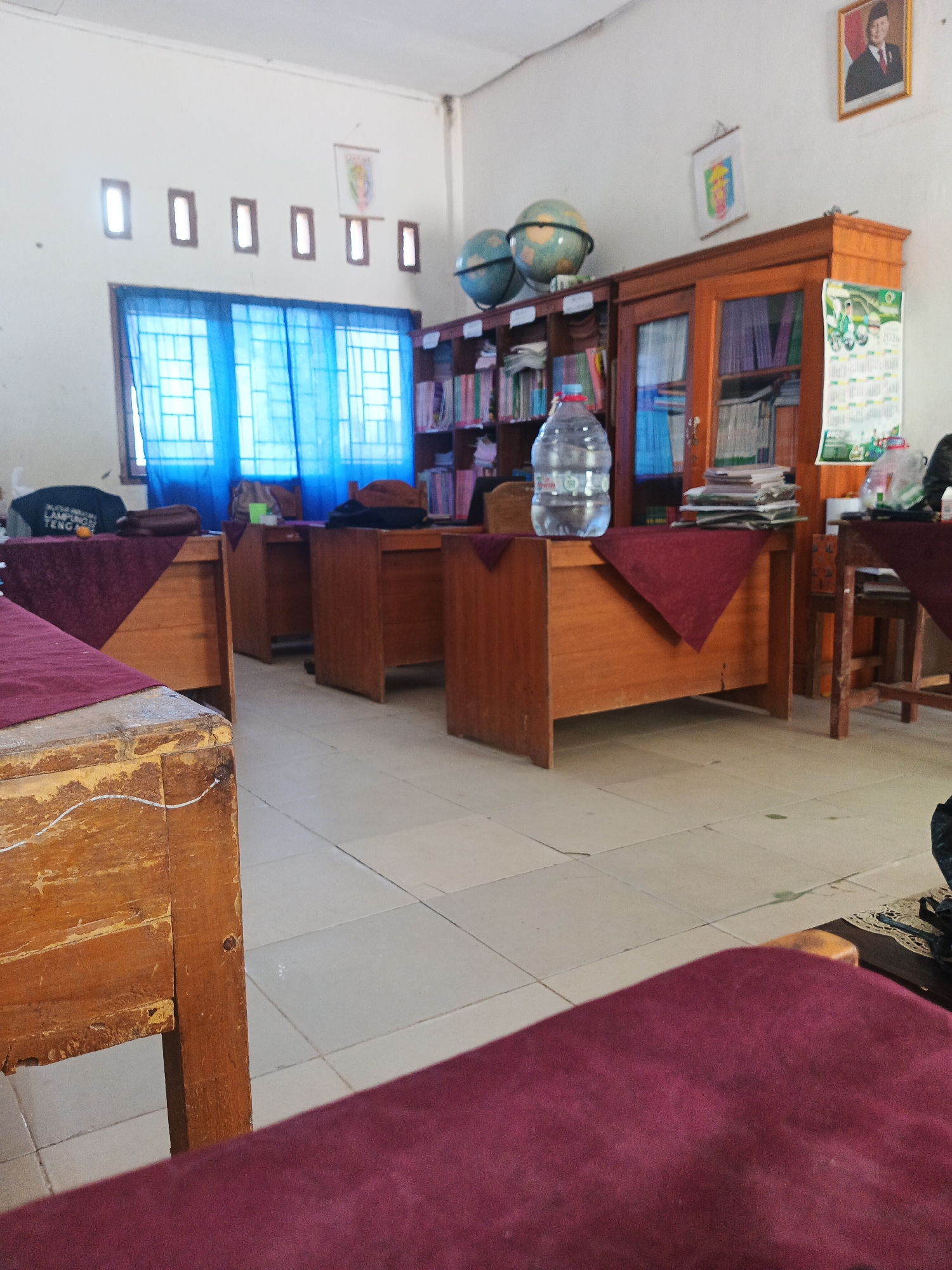 The image depicts a classroom or office environment, likely in Rejo Basuki, Indonesia, with a focus on the interior details. In the foreground, the edge of a wooden table or desk is visible, covered with a dark red fabric. Beyond this, several similar tables are arranged in rows, also draped with the same red fabric. These tables appear to be individual workstations.
Further into the room, a large wooden cabinet with glass doors stands against the wall. The cabinet contains shelves filled with books and what appear to be files or folders. Two globes are positioned on top of the cabinet. Above the cabinet, a framed portrait of a man in a suit and tie is displayed on the wall, alongside a smaller framed crest or emblem.
To the left of the cabinet, a window covered by bright blue curtains allows light to enter the room. Above the curtains, a series of small, rectangular openings are visible in the wall, possibly for ventilation. A large plastic bottle of water is placed on one of the tables, near the cabinet. The floor is tiled in a light color. The overall impression is of a functional, perhaps slightly dated, educational or administrative space. There are no visible people in the image.