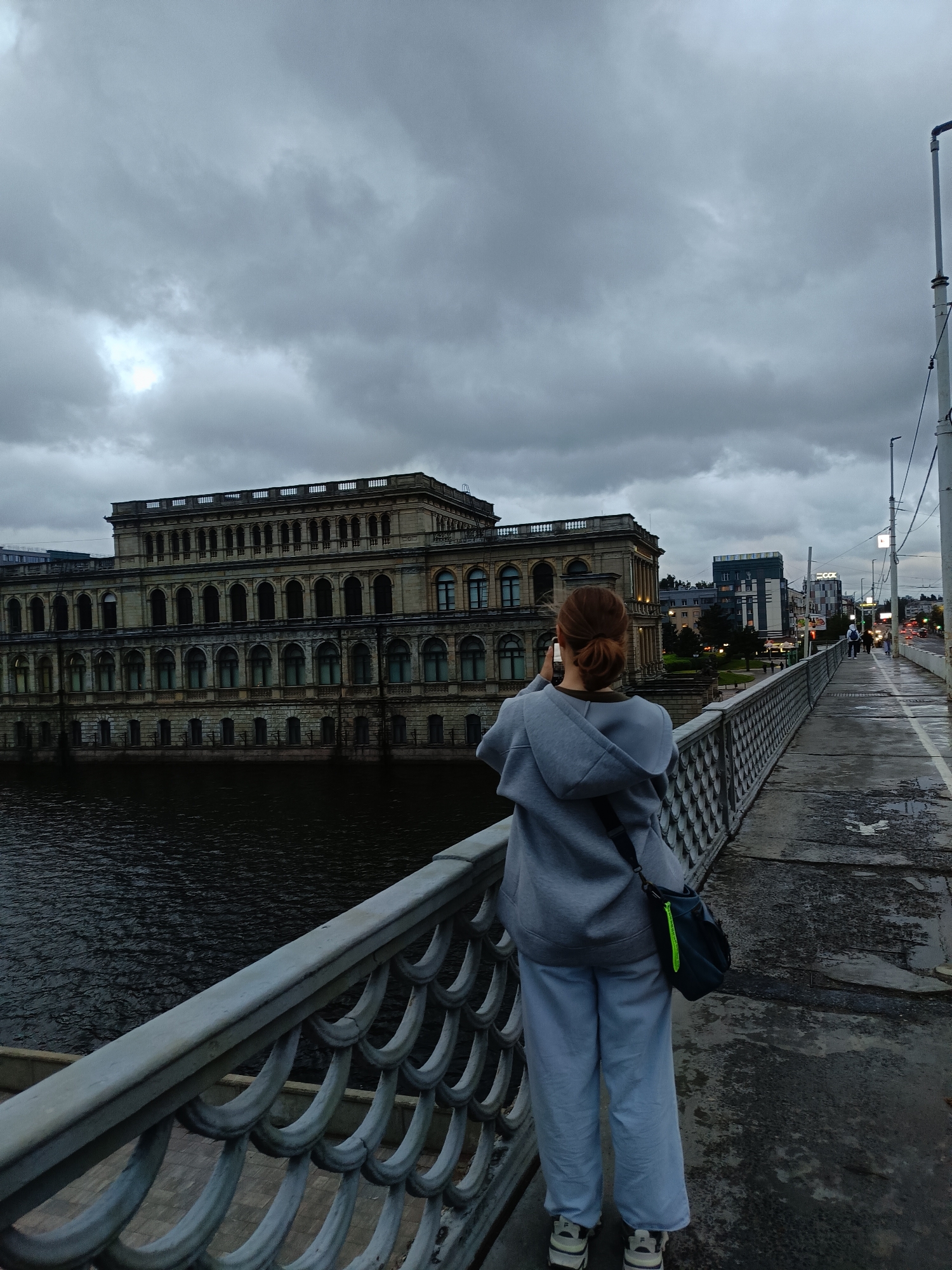 A person is standing on a bridge, facing away from the camera, and taking a photo with their phone. They are wearing a grey hoodie, grey pants, and sneakers. A black bag with a neon green zipper hangs from their shoulder. The bridge has a decorative metal railing. To the left of the bridge is a body of dark water. Across the water is a large, ornate, multi-story building with many arched windows. Beyond this building, the city skyline is visible, with modern buildings and streetlights. The sky is overcast and grey, with a patch of brighter light suggesting the sun trying to break through. The bridge's walkway appears wet, with puddles visible, indicating recent rain. The location is the Upper Pond embankment in Kaliningrad, Russia, with the iconic "House of Soviets" building visible in the background.