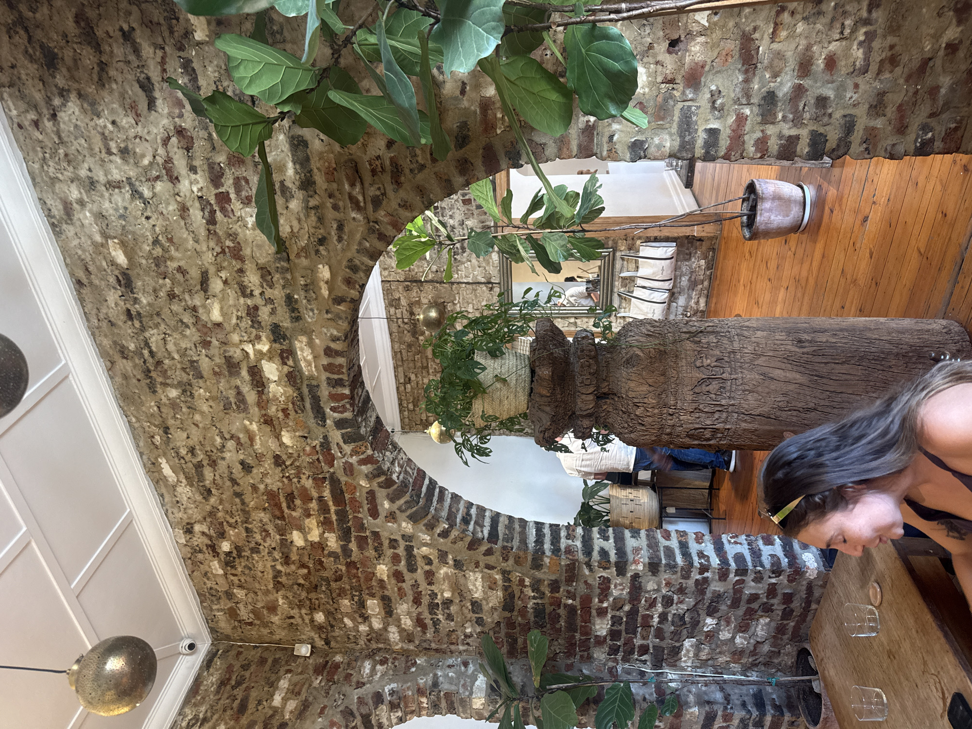 A vertically-oriented interior shot presents a restaurant setting, evidenced by a woman seated at a wooden table, seemingly interacting with glassware on it. Her presence is partially obscured by a large, sculpted wood piece, likely a table leg or decorative element. The scene is set within a space characterized by exposed brick archways. Above, a glimpse of a white-paneled ceiling can be seen, while in the background, through the archway, there is additional seating with white seat cushions and a mirror. There are various green plants throughout the scene. The setting has been identified as Cape Town, South Africa.