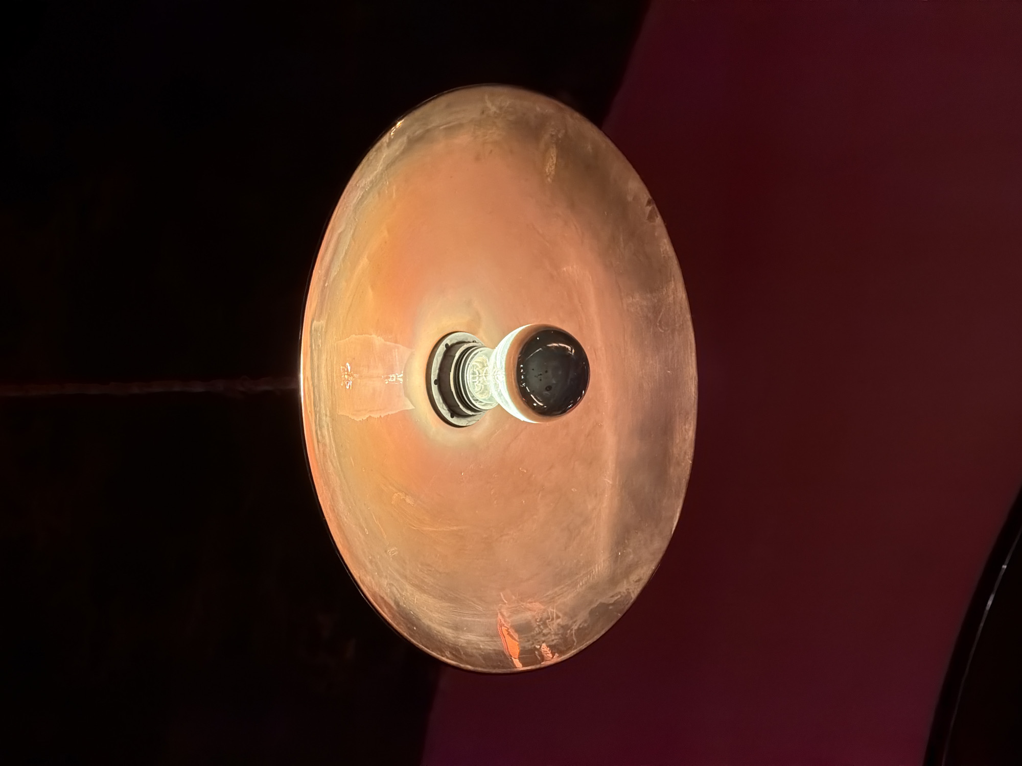 The image shows a close-up, low-angle shot of a single, vintage-style incandescent light bulb hanging from a black cord. The bulb is enclosed within a large, shallow, translucent lampshade with an orange-amber hue, creating a warm, diffused light. The lampshade appears to be made of glass with some subtle imperfections or texture. The background consists of a dark, possibly black, ceiling above the lampshade and a deep red wall below. The overall impression is one of dim, atmospheric lighting. The image does not provide any information about a specific location in Milan, Italy, or any people, actions, or interactions.