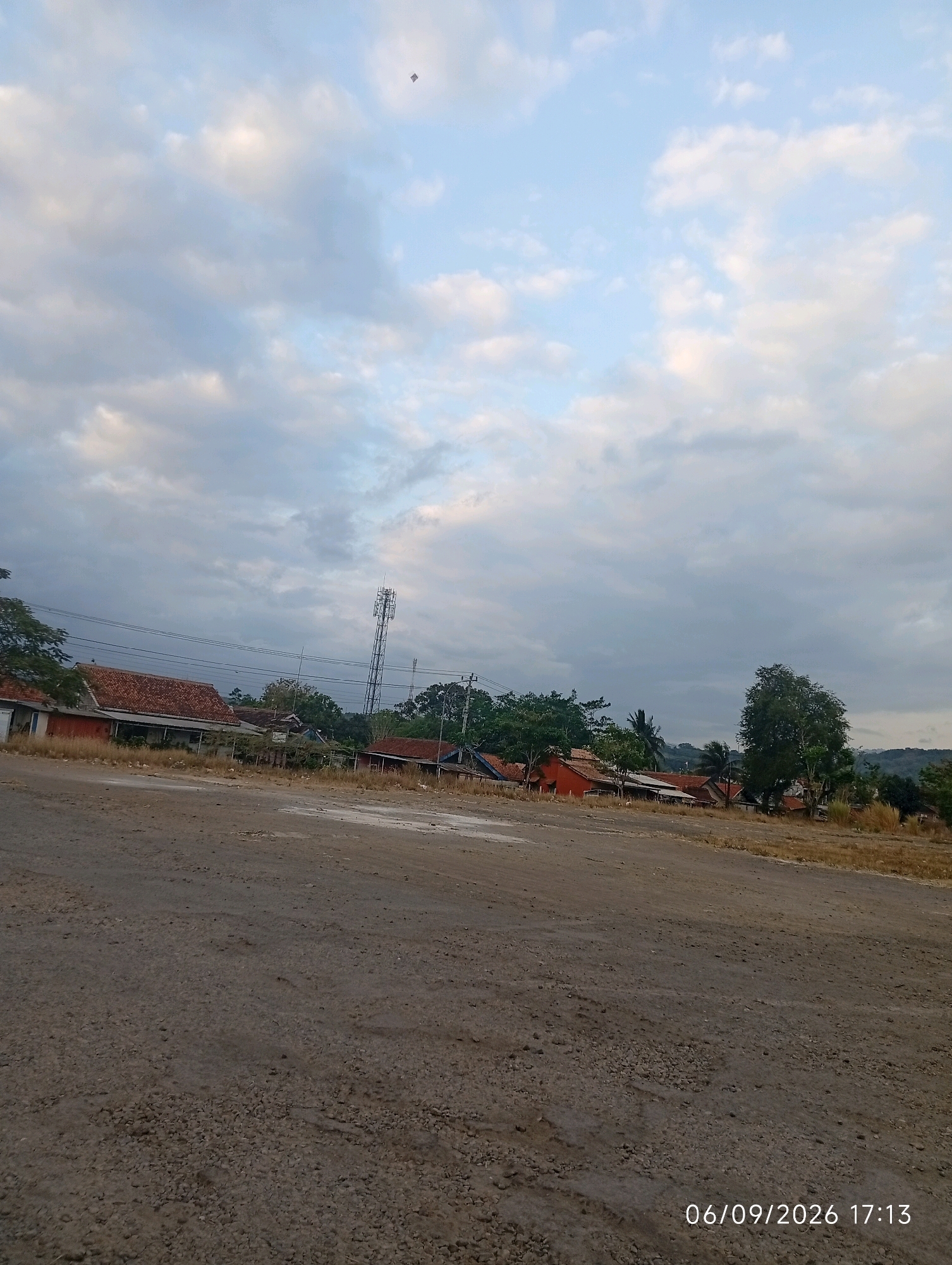 An aerial view of a dirt road in the city of Purworejo, Indonesia. The road is covered with gravel and dirt. In the distance, there are houses with red tile roofs, a cell tower, and trees. The sky is cloudy with a hint of blue peeking through. There is a small kite flying in the sky. The date and time stamp on the image reads "06/09/2026 17:13".