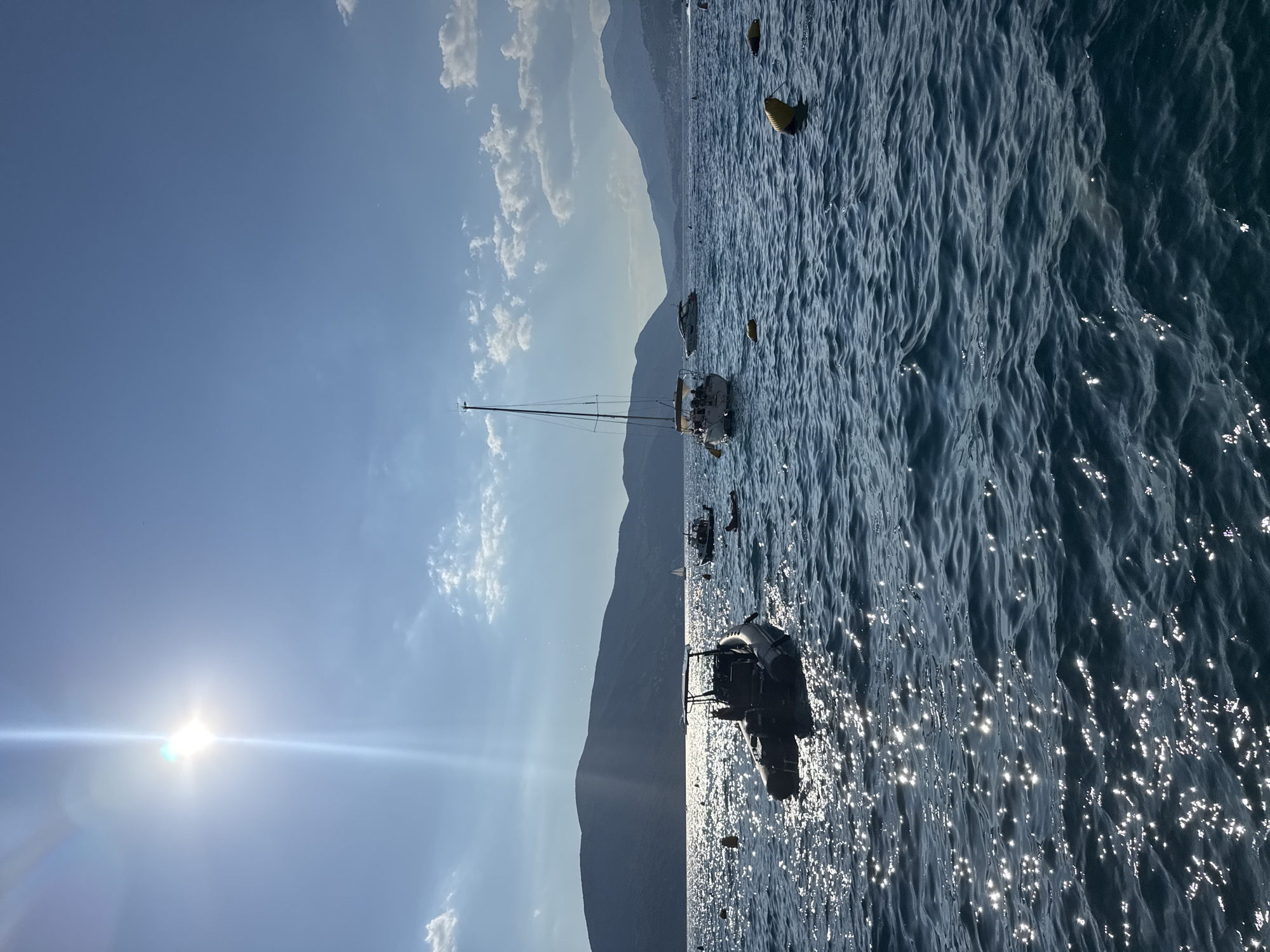 This is an outdoor, high-angle, full shot of a body of water with several boats and mountains in the background. The sun is shining brightly, creating a glare on the water and a lens flare effect on the left side of the image. The sky is a clear blue with a few scattered white clouds. The mountains are dark and appear to be covered in vegetation.
There are several boats on the water. In the foreground, there is a rigid inflatable boat with a console and seating. Further out, there is a sailboat with its mast visible. In the distance, there are other smaller boats and buoys. The water is choppy with small waves, and the sunlight reflects off the surface, creating a shimmering effect.
The time of day appears to be late morning or early afternoon, given the position of the sun and the quality of light. The weather is clear and sunny. The scene evokes a sense of peace and tranquility, suggesting a recreational outing on the water. There is no visible text in the image.