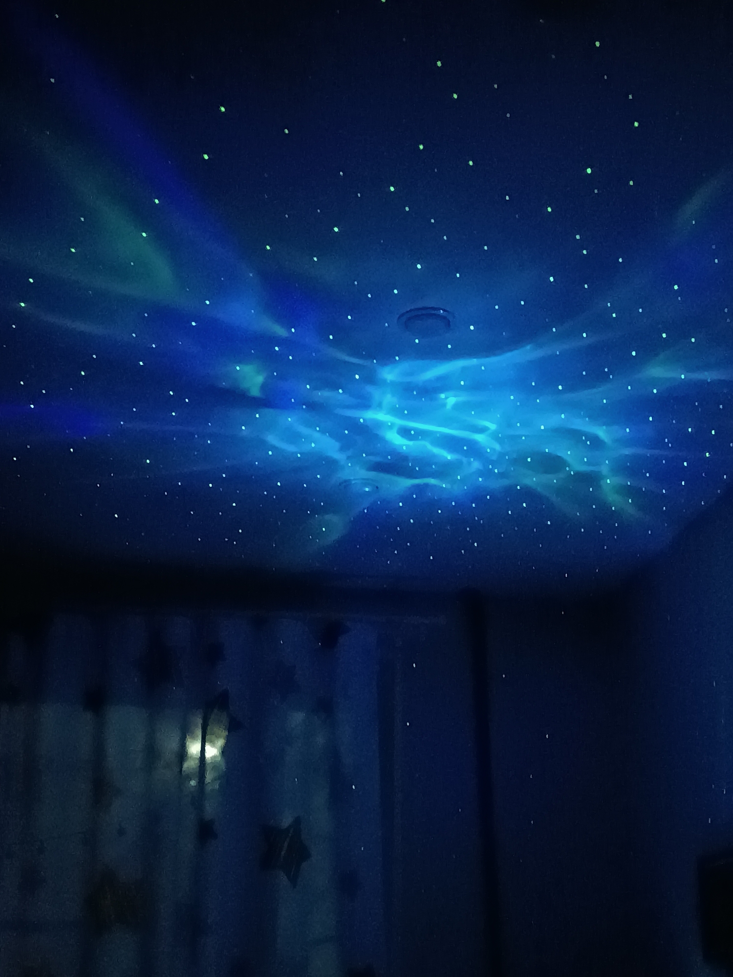 This image depicts a dark indoor room with its ceiling transformed into a starry night sky. Numerous small, bright green "stars" are scattered across the dark blue ceiling. Dynamic, swirling patterns of blue and light teal light are projected onto the ceiling, resembling nebulae, aurora borealis, or water reflections, creating an ethereal and fluid visual effect. A dark, circular fixture, likely a vent or recessed light, is visible on the ceiling towards the left side.
The room is dark, suggesting it's either nighttime or purposefully dimmed to enhance the light projection. On the left side of the image, a window is partially visible, covered by a sheer curtain through which faint, dark star-shaped decorations can be discerned. The lower right portion of the image shows a dark, indistinct area, possibly furniture or another part of the room. No people are present, and no activity is explicitly occurring beyond the visual light display. The scene evokes a sense of calm, wonder, and a dreamlike atmosphere, likely intended for relaxation or as a decorative element in a bedroom. There is no visible text.