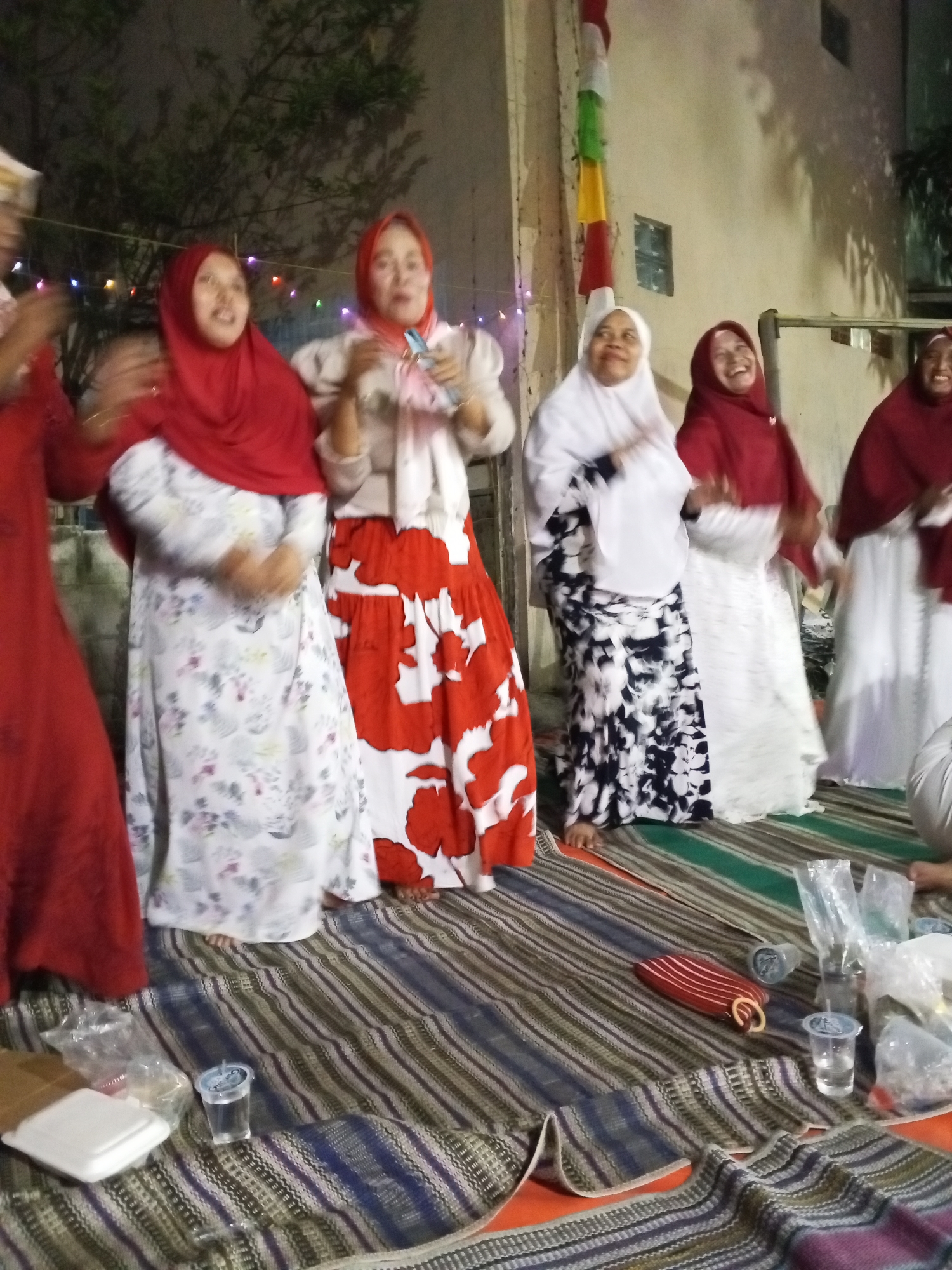 Six women are gathered outdoors in what appears to be a residential area in Ciruas, Indonesia. The women are all wearing headscarves, with four of them wearing hijabs. They are standing on a striped woven mat, with some seated on the mat as well. The women are dressed in long, loose-fitting garments. Two of the women are holding items, one in her right hand and the other in her left. One woman is laughing heartily, with her head tilted back. In the foreground, there are several plastic cups with clear liquid, as well as plastic bags and a small red and white striped object with a ring. The background shows a building wall, a tree, and decorative lights strung across the scene. The overall impression is of a social gathering, likely a celebration or informal get-together.