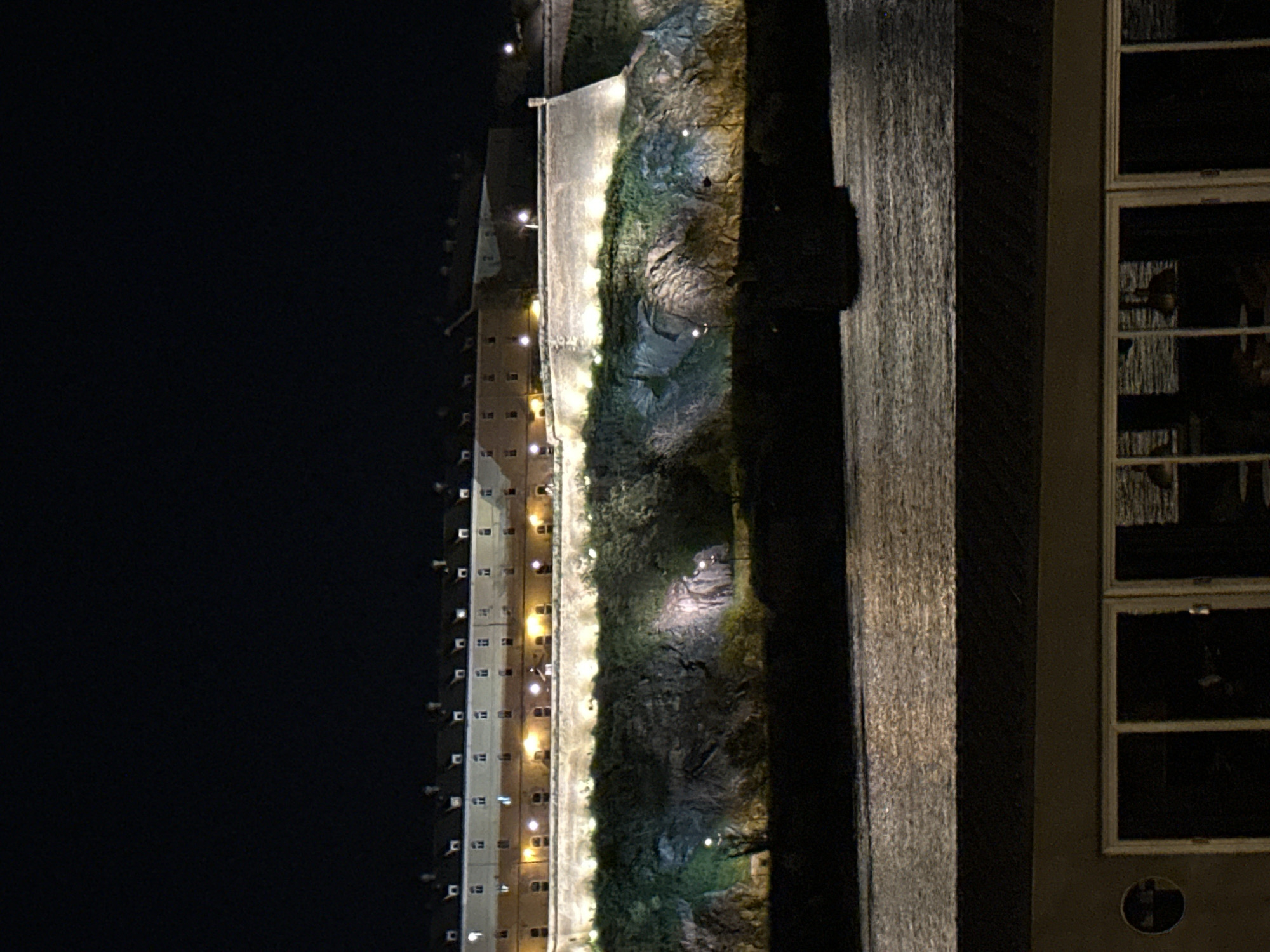 Captured at night, the scene depicts a building facade in Novi Sad, Serbia. A tall, slender building is illuminated with rows of lights lining its height. Next to it, a textured wall simulates a rock face, also lit with sparse lights. Adjacent to the rock wall is a smooth, dark, possibly wooden structure. Further right, a section of a building with windows is visible. The overall impression is urban architecture with design elements incorporated into the building's facade.