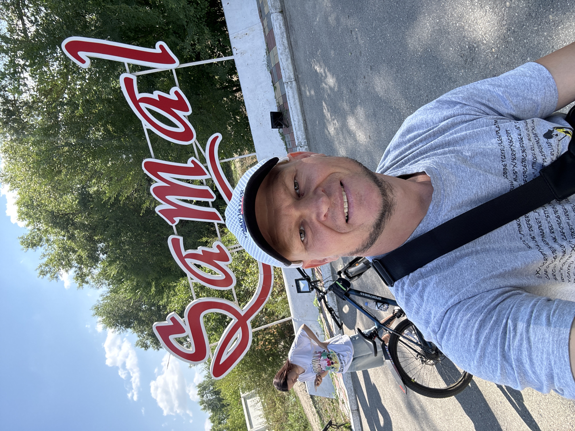 In Oskemen, Kazakhstan, a man is taking a selfie in front of a large sign that reads "Samal 1." He is wearing a grey t-shirt and a white and blue checkered baseball cap. Behind him, a woman in a white t-shirt and denim shorts is looking at her phone, standing next to a bicycle. Another bicycle is visible to the left. The background features lush green trees under a clear blue sky with scattered clouds. The sign is red with white trim and mounted on a white structure. The ground is asphalt, and there is a paved walkway with decorative curbing.