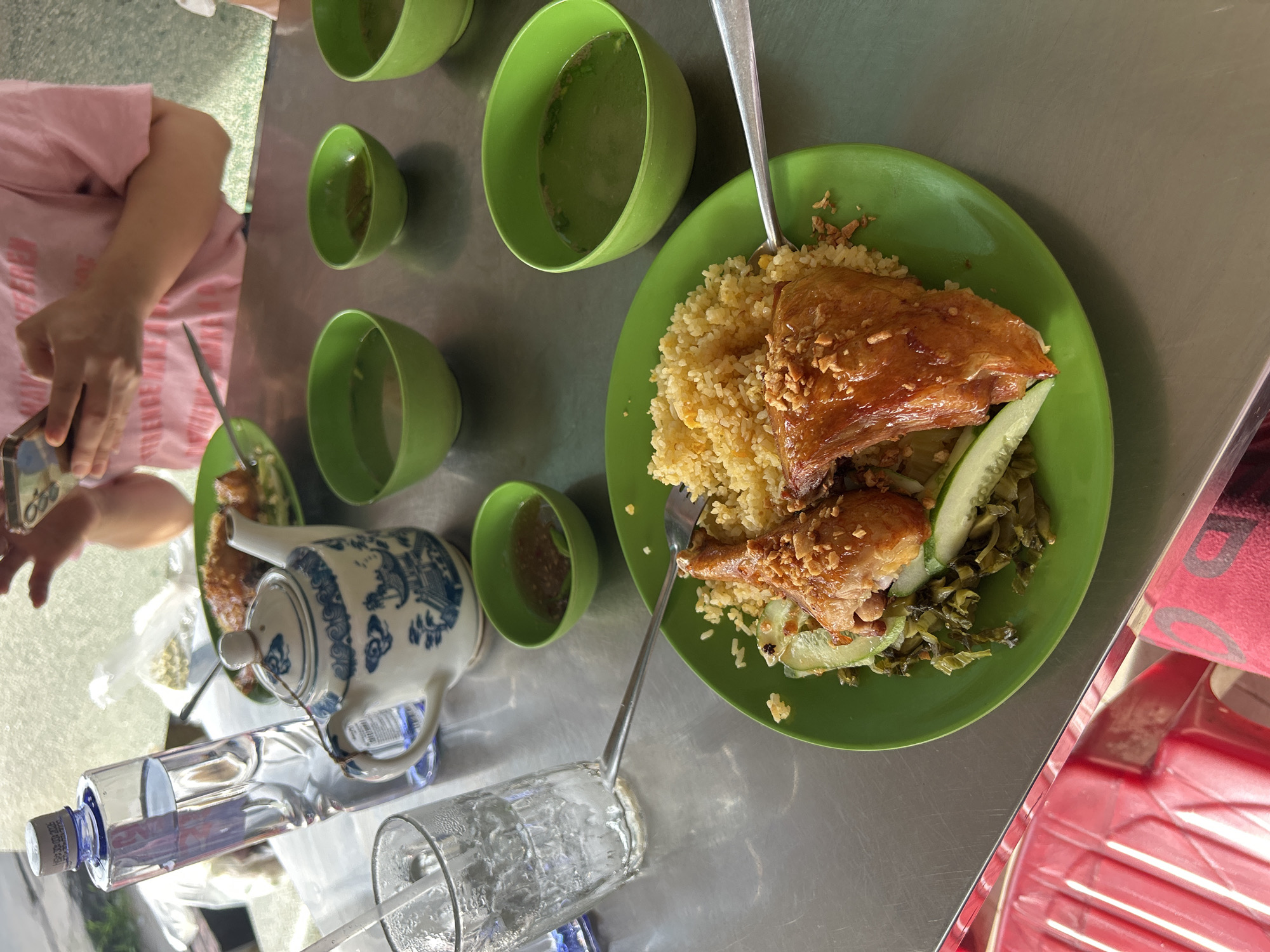 In An Hữu, Vietnam, a close-up shot reveals a meal laid out on a metallic table. A vibrant green plate holds a portion of glistening rice topped with chopped nuts and a piece of glazed chicken, accompanied by sliced cucumber and pickled greens. To the right, three small green bowls contain a light-colored broth. A clear glass with ice and a straw sits to the left, next to a decorative blue and white teapot. Behind these items, a tall bottle of clear liquid stands upright, its cap secured. In the upper background, a person in a pink t-shirt is visible, holding a mobile phone. The scene suggests a casual dining experience in a shaded area, possibly outdoors due to the lighting.