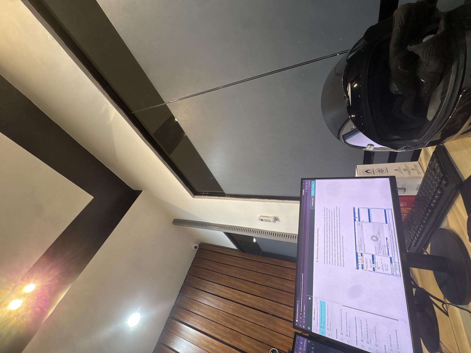 The image shows a desk with a computer monitor displaying an online course about remarketing personal lines policies. The course content is visible on the screen, along with a navigation sidebar. A black motorcycle helmet is placed on the desk, partially obscuring the keyboard. The background features a wooden-paneled wall and a modern ceiling with dark and light accents. Lights are visible on the ceiling. A security camera is mounted on the wooden wall. The scene appears to be an indoor workspace, possibly an office or home office, located in Davao City, Philippines.
