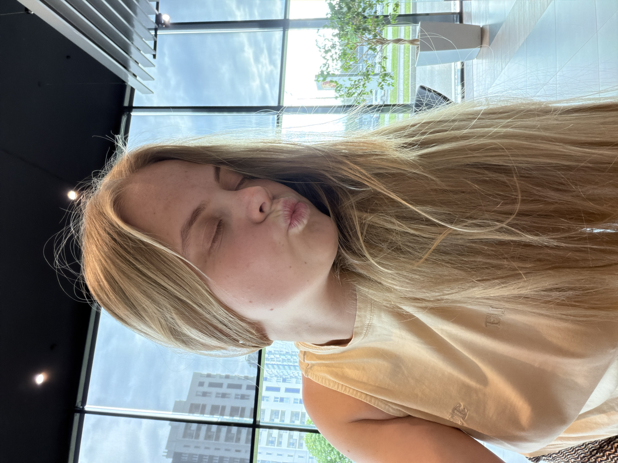 A fair-skinned young woman with long blonde hair is oriented sideways in the frame, puckering her lips for a kiss. She wears a light beige t-shirt. Behind her, through large windows, a cityscape is visible, potentially in Izhevsk, Russia. Details include buildings with multiple windows, and a tree. The interior includes a dark ceiling with lights and possible air vents.
