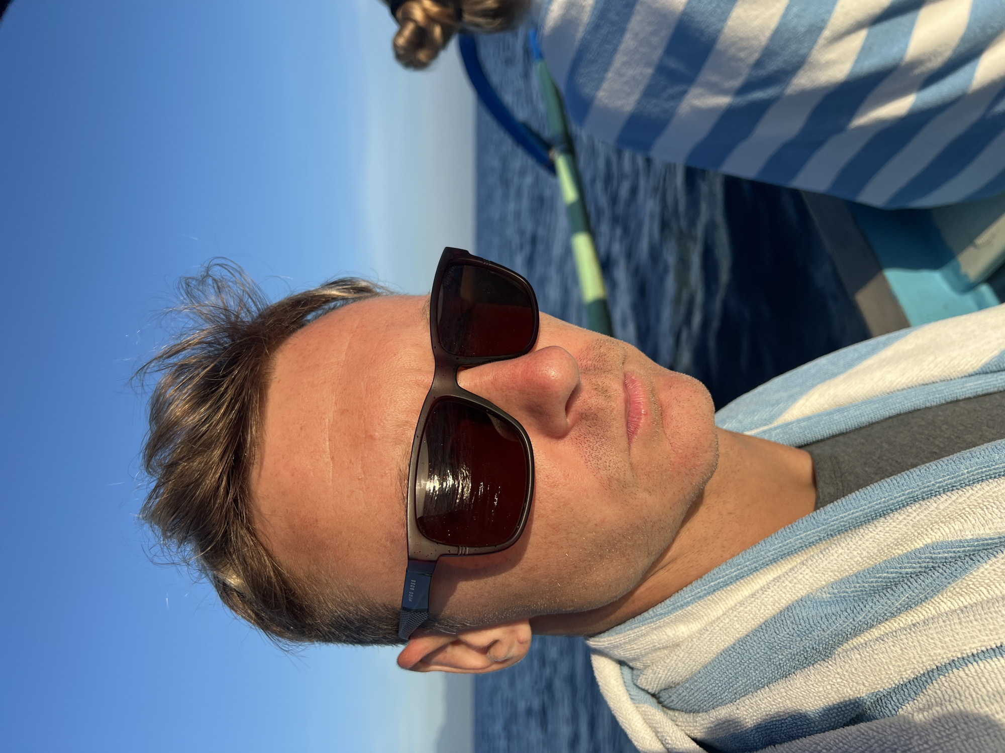 A medium shot shows a man wearing sunglasses and a blue and white striped towel on a boat. The background includes a body of water and a blue sky. The location is Seririt, Indonesia.