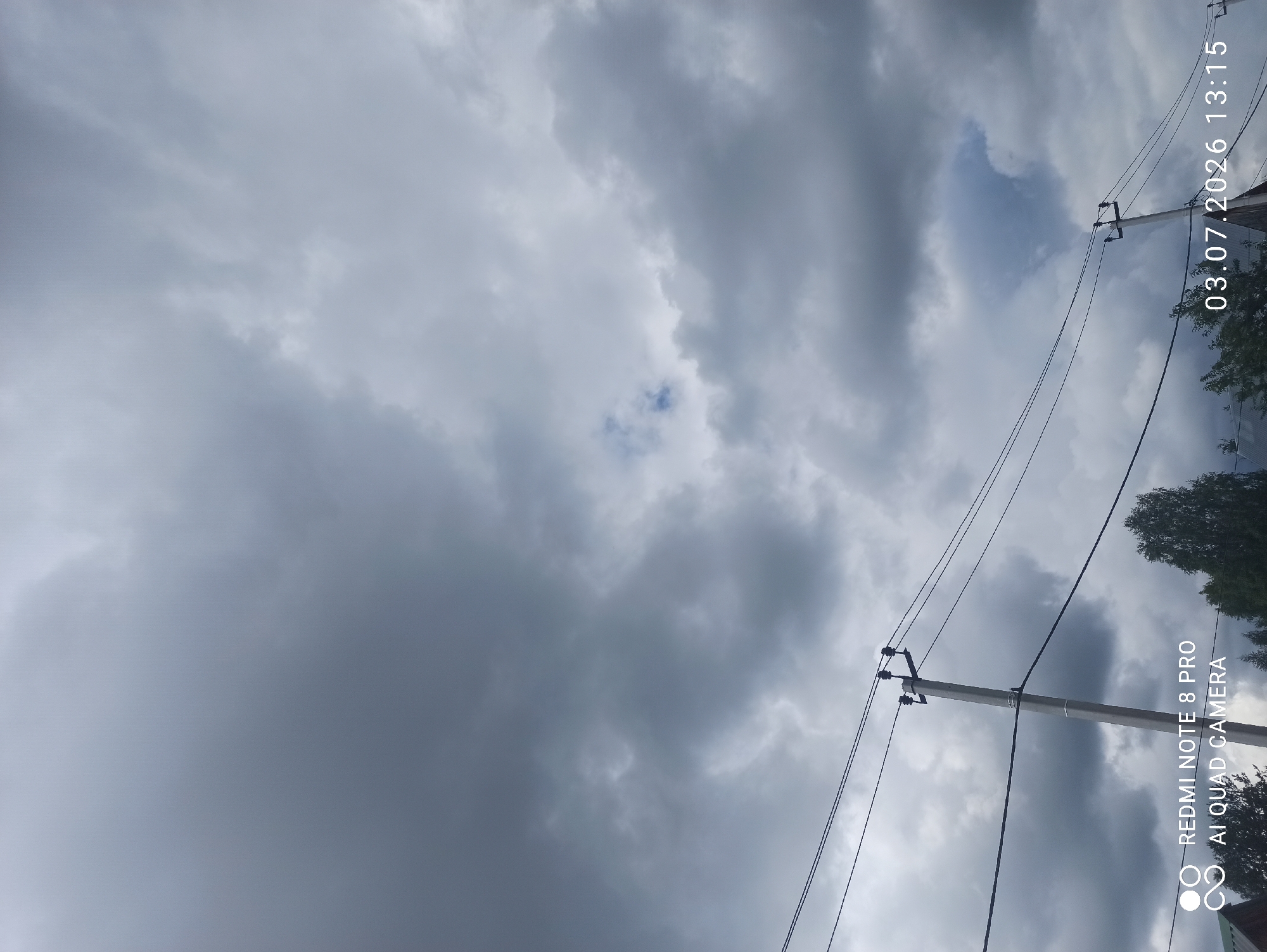 An outdoor, medium shot photograph depicts a cloudy sky with power lines and utility poles in the foreground. The sky is filled with dark, heavy clouds, suggesting overcast or stormy weather. Two utility poles are visible, with several power lines strung between them. The poles are grey and appear to be made of concrete. Below the power lines, the tops of trees and the roof of a building can be seen. A timestamp in the lower right corner indicates the date and time as "03.07.2026 13:15". In the lower left corner, a watermark displays "REDMI NOTE 8 PRO AI QUAD CAMERA". The image was taken in the city of Sovetskiy, Russia. There are no people or animals visible in the frame.