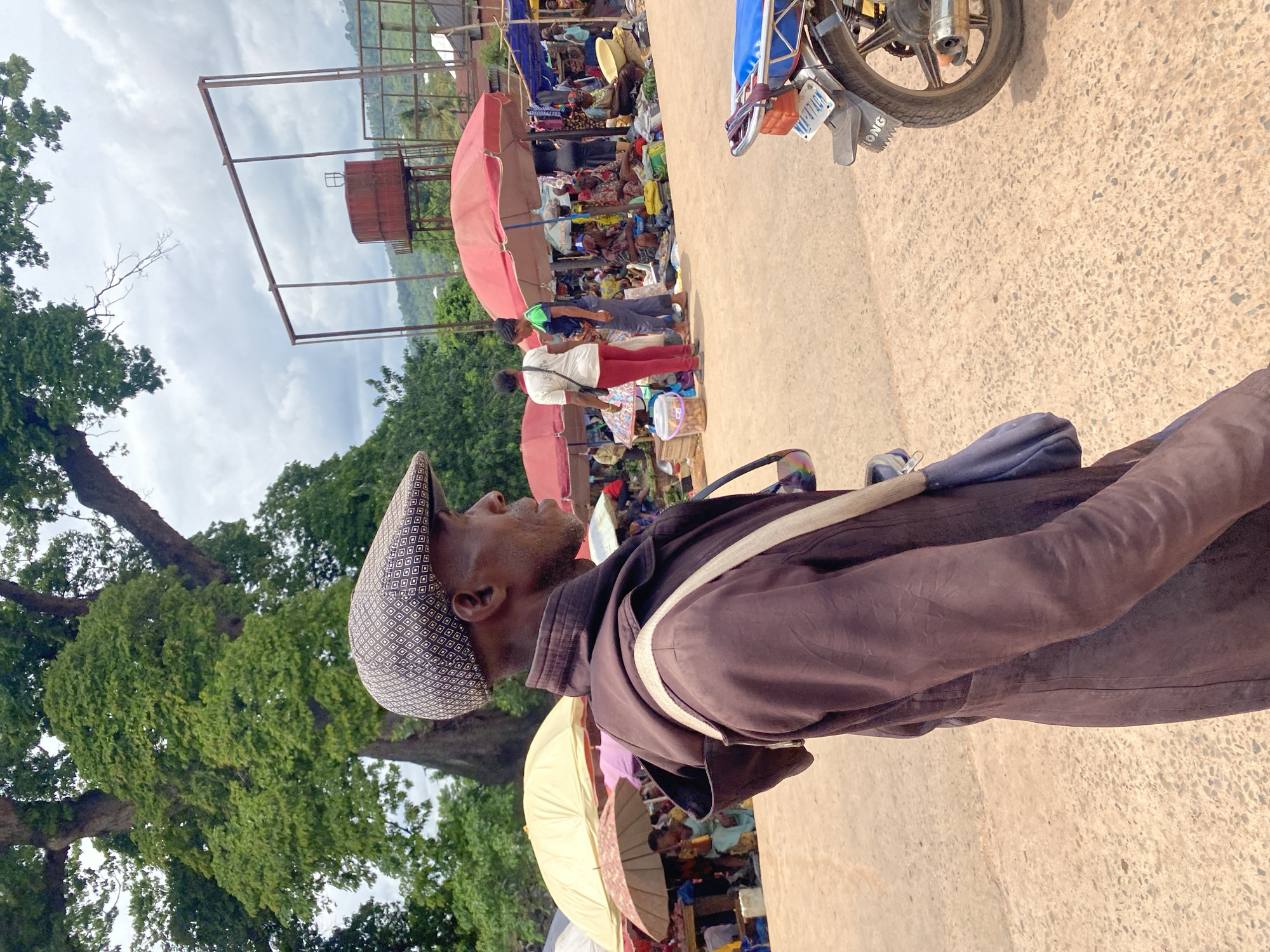 The image shows a man standing in an open market in Igbara, Nigeria. The man is wearing a patterned cap and a brown jacket, with a bag slung across his shoulder. The market has many people selling under red and white umbrellas. A motorcycle is parked nearby. The sky is cloudy, and there are trees in the background. A rusty metal tower stands in the distance.