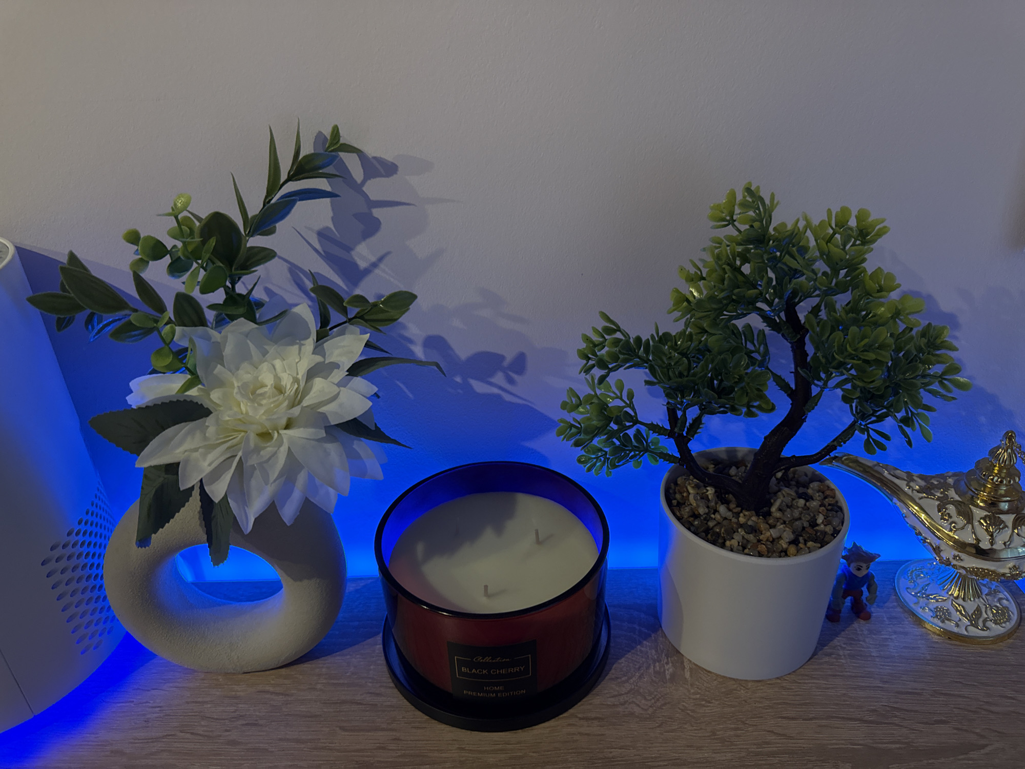 A still life composition rests on a light brown surface, possibly a wooden shelf. From left to right: a cylindrical object with vertical slits emitting blue light, a white donut-shaped vase containing a white flower arrangement, a dark red candle with a "Black Cherry" label, a small potted artificial bonsai tree with pebbles, a small plastic figurine of a person, and a white and gold genie lamp. The backdrop is a pale wall. The lighting is predominantly blue, possibly from an LED strip. The location in the city of Lovech, Bulgaria is not evident in the scene.