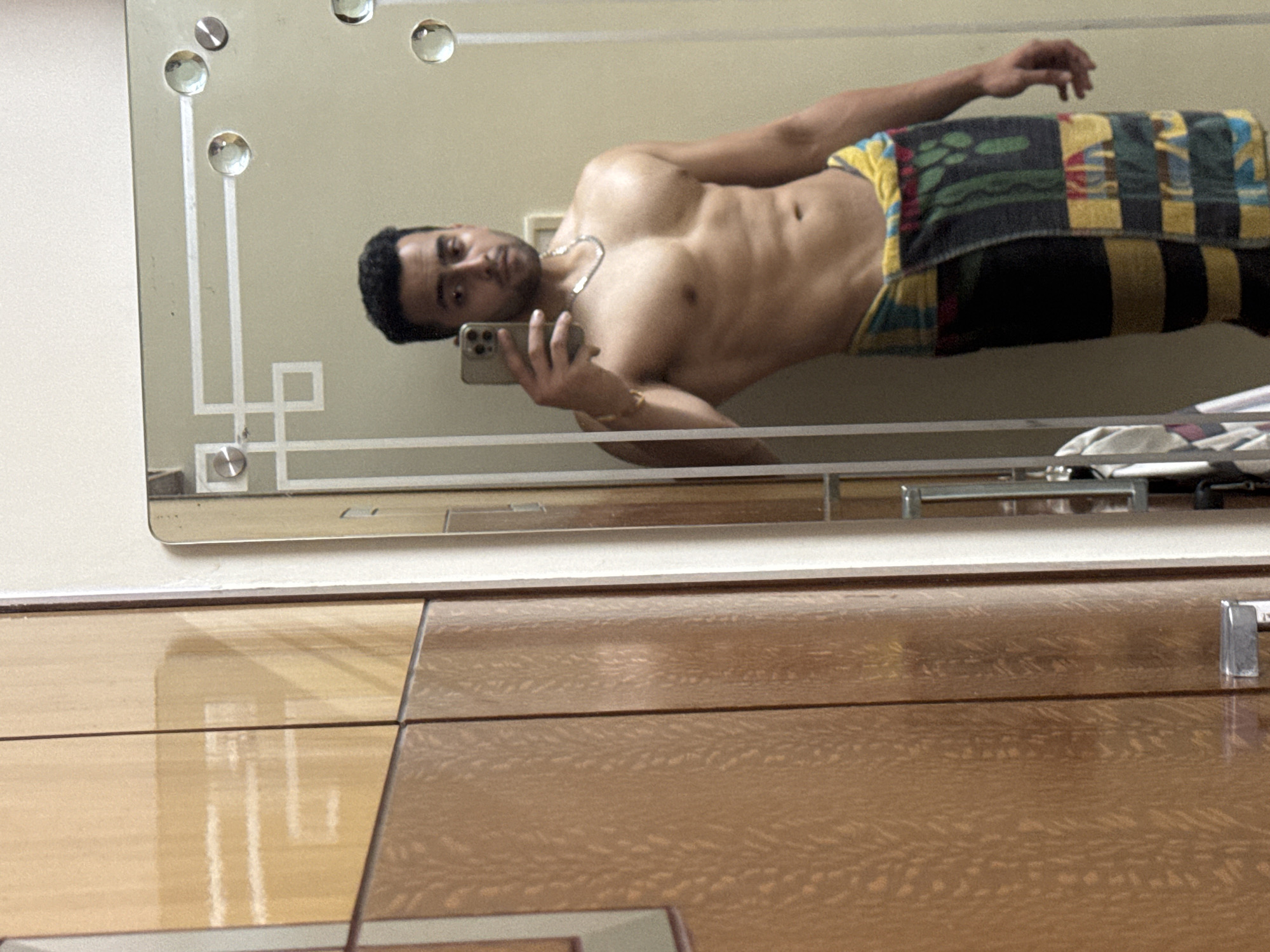 A shirtless man is taking a selfie in a mirror. He is wearing a colorful towel around his waist and has a gold chain around his neck. The mirror is mounted above a dresser with a glossy finish. The location is unknown and there is no explicit connection to the city of Kolkata, India.