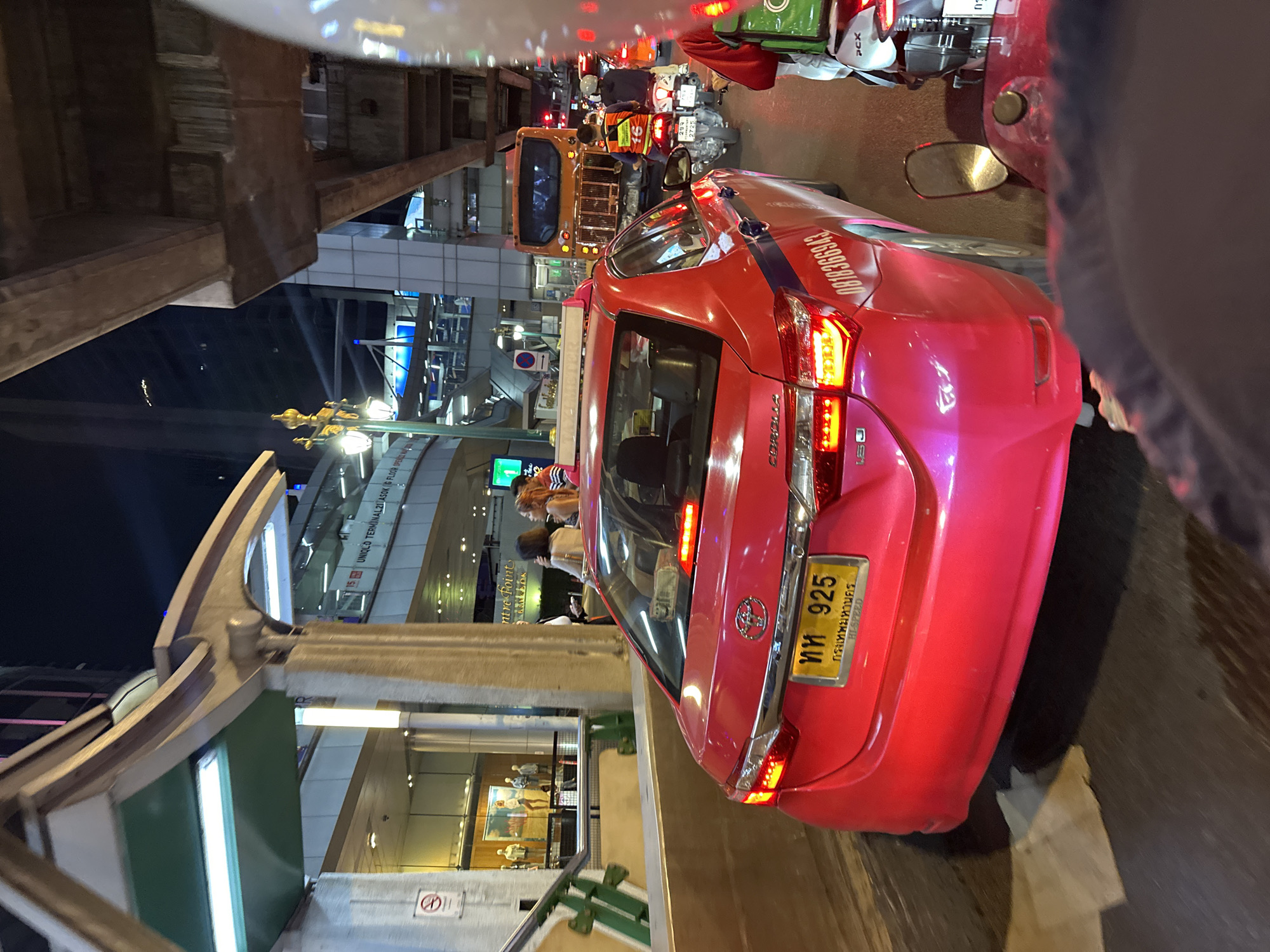 The image depicts a traffic scene in Bangkok, Thailand, at night. A pink Toyota Corolla taxi is stuck in heavy traffic underneath a pedestrian overpass. The license plate number is ทห 925, registered in Bangkok. People are walking on the pedestrian overpass, which leads to a shopping mall, specifically Terminal 21 Asok. Various vehicles, including motorcycles and a yellow tuk-tuk, are visible in the congested street. The surroundings are illuminated by streetlights and building lights. A sign indicates that the UNIQLO store is located on the G floor of the Terminal 21 Asok shopping mall.