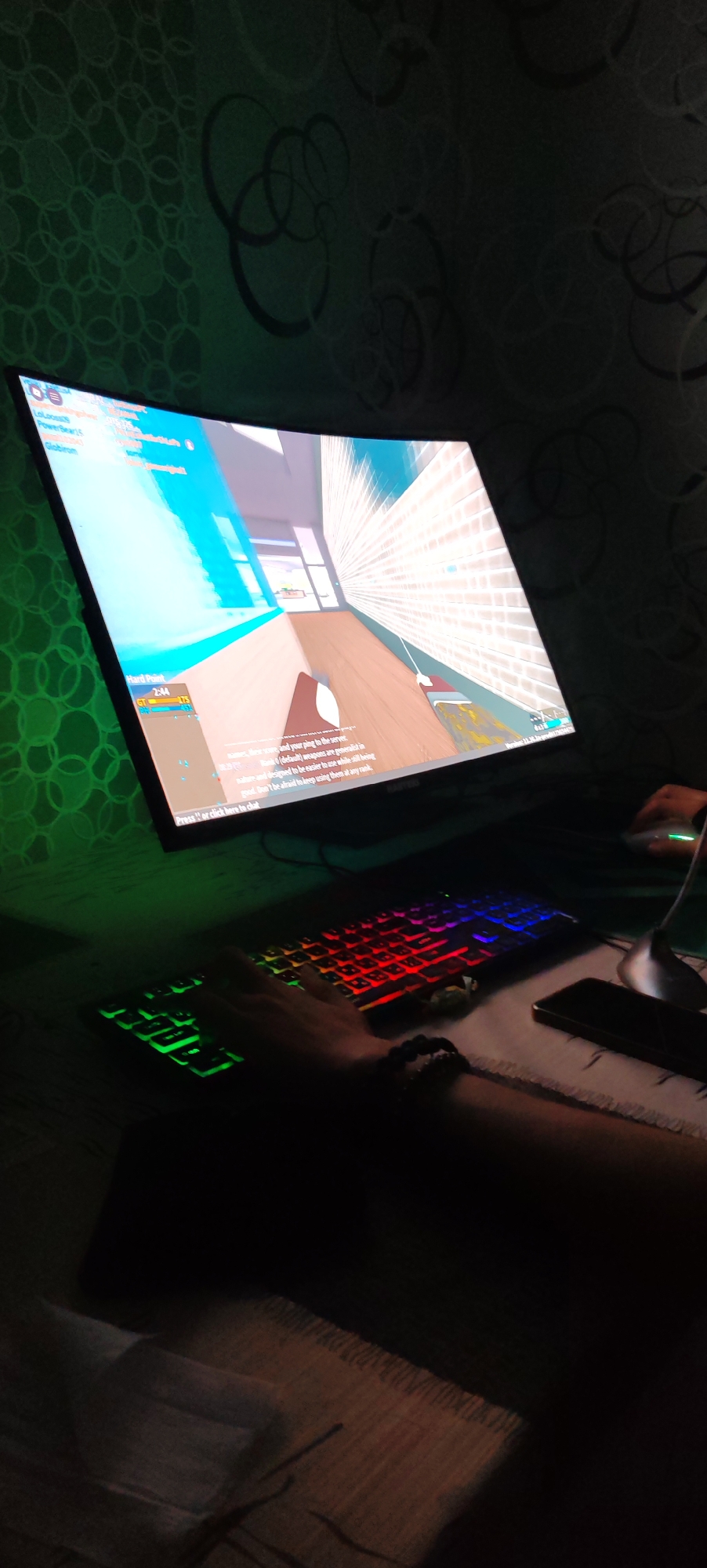 An indoor, eye-level, medium shot captures a person's hands playing a video game on a computer. The computer monitor, which takes up the left half of the frame, displays a first-person perspective game with bright, cartoonish graphics. A colorful, RGB-backlit keyboard is visible in the lower half of the frame, with the player's left hand resting on it. A mobile phone and a computer mouse are also on the table. The background is a dark, textured wallpaper. The lighting is dim and primarily comes from the computer screen and keyboard, casting a green glow on the wall. The image suggests a gaming session taking place in a private residence. The location is not specified in the image.