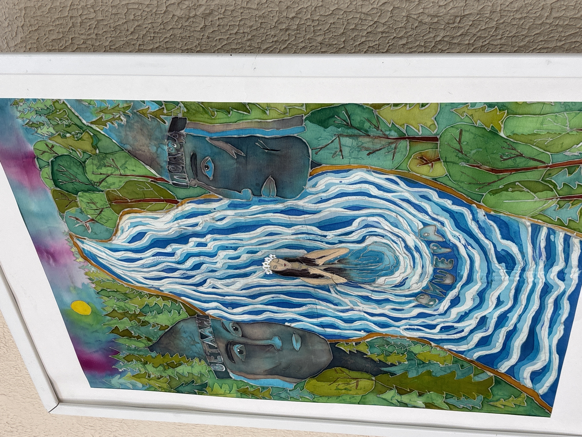 The image shows a framed piece of artwork depicting a river scene. A woman with dark hair and a flower crown floats in the swirling blue and white water. On either side of the river are two large, stylized faces embedded in the green foliage, suggesting river spirits or guardians. Above one face, the name "BETAHAH" is written. A yellow sun is visible on the left side of the artwork. The artwork is framed in white and is resting on a surface with a textured beige wall behind it. This artwork is located in the city of Chaikovsky, Russia.