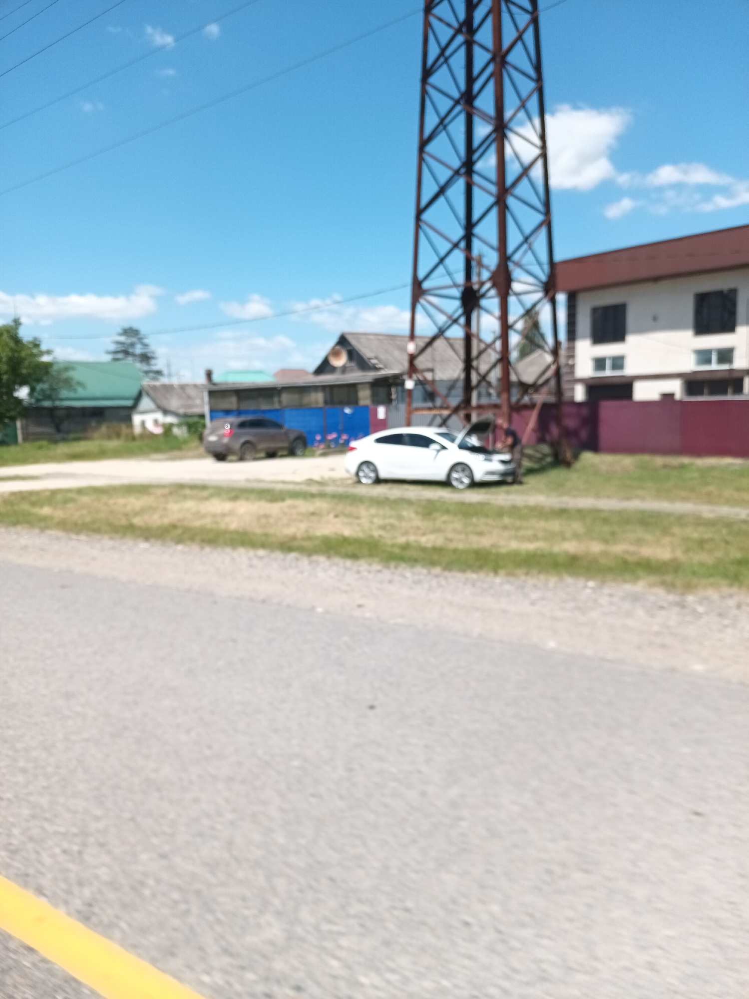 The image shows a residential area with houses, a power line tower, and two cars on a sunny day. A brown sedan is parked on the left side of the frame, and a white sedan with its hood open is parked on the right, with a person visible near the open hood. The ground is a mix of gravel and grass. The sky is blue with scattered clouds. This scene is located in the city of Krymsk, Russia.