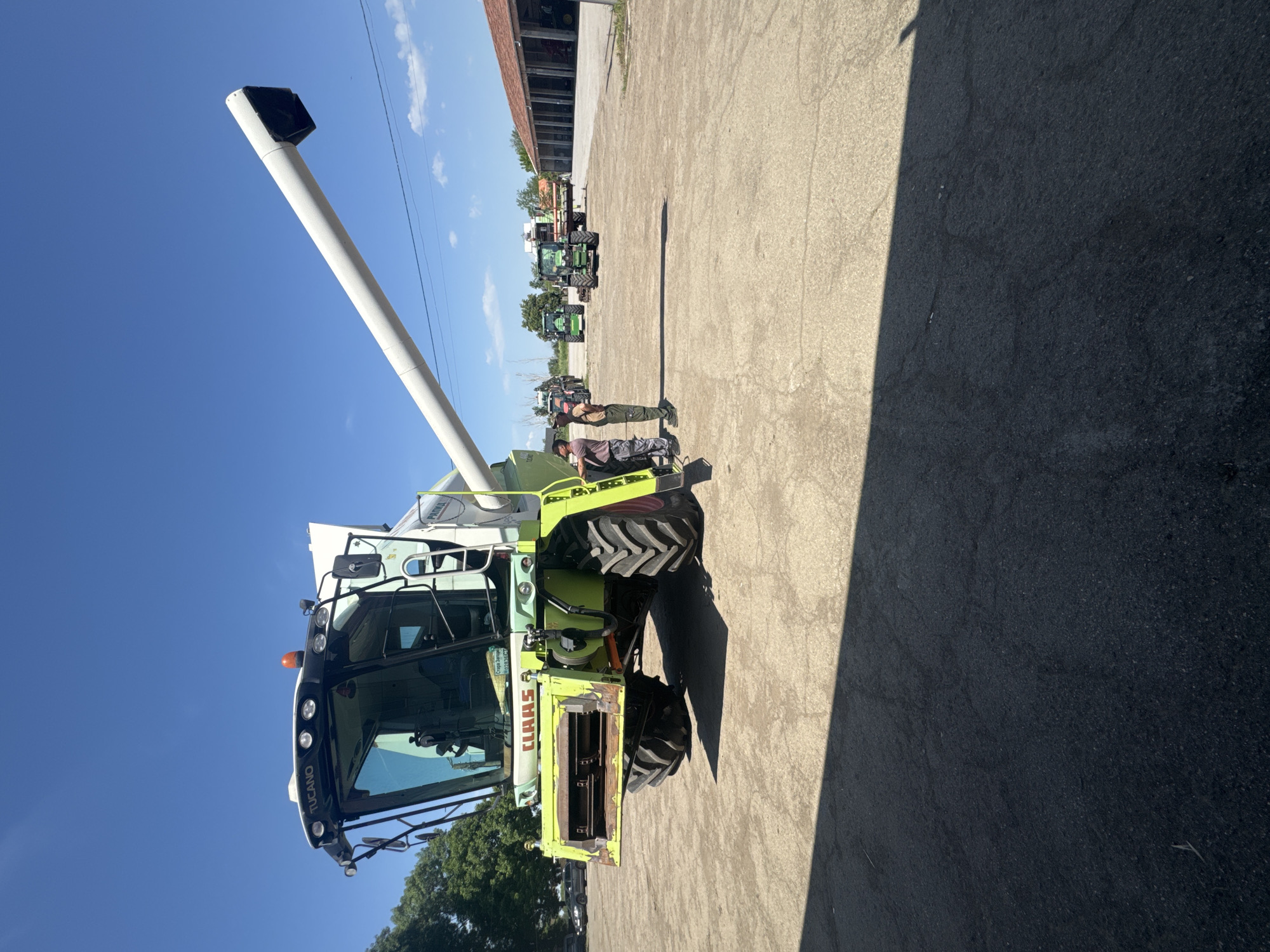 The image depicts a green and white Claas Tucano combine harvester parked on a concrete surface under a clear blue sky. Two individuals are standing near the combine. In the background, there are several green tractors and a building. The location is Sarnevo, Bulgaria.