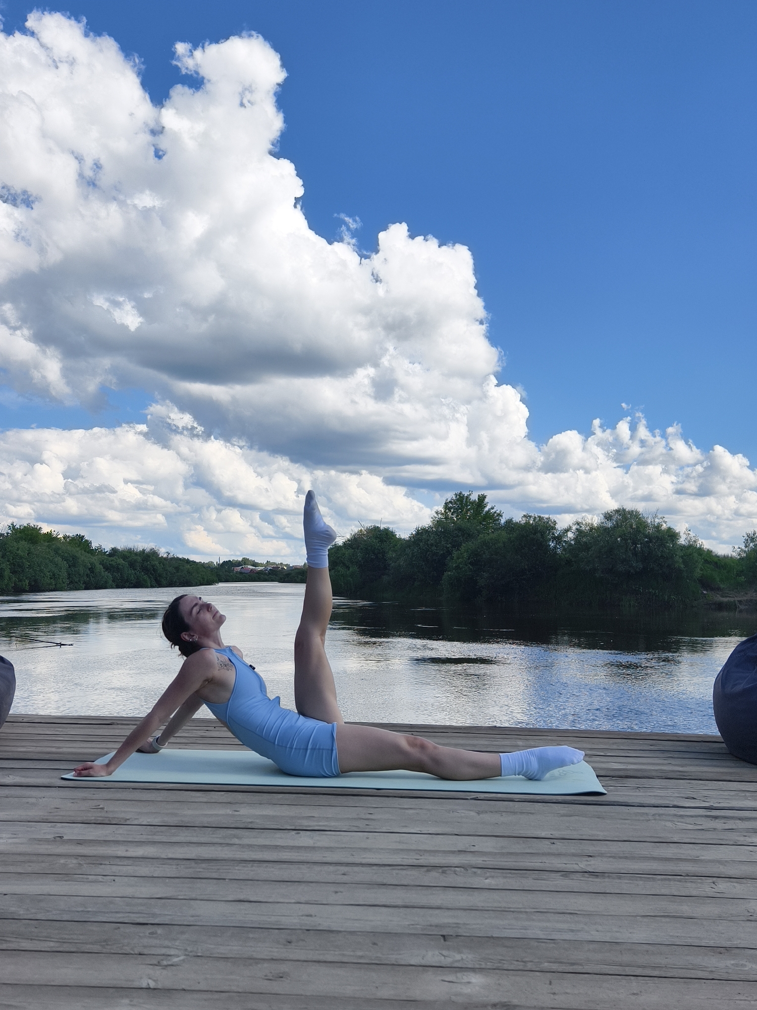 A woman is performing a yoga pose on a light blue yoga mat on a wooden pier. She is lying on her back with her legs extended, one leg raised vertically and the other bent at the knee. She is wearing a light blue leotard and white socks. Her eyes are closed, and her head is tilted back. The pier is located next to a body of water, likely a river, with a line of trees and vegetation on the opposite bank. The sky is blue with scattered white clouds. The scene appears to be outdoors, with no visible buildings or structures belonging to the city of Онохино, Russia, other than possibly in the distant background.