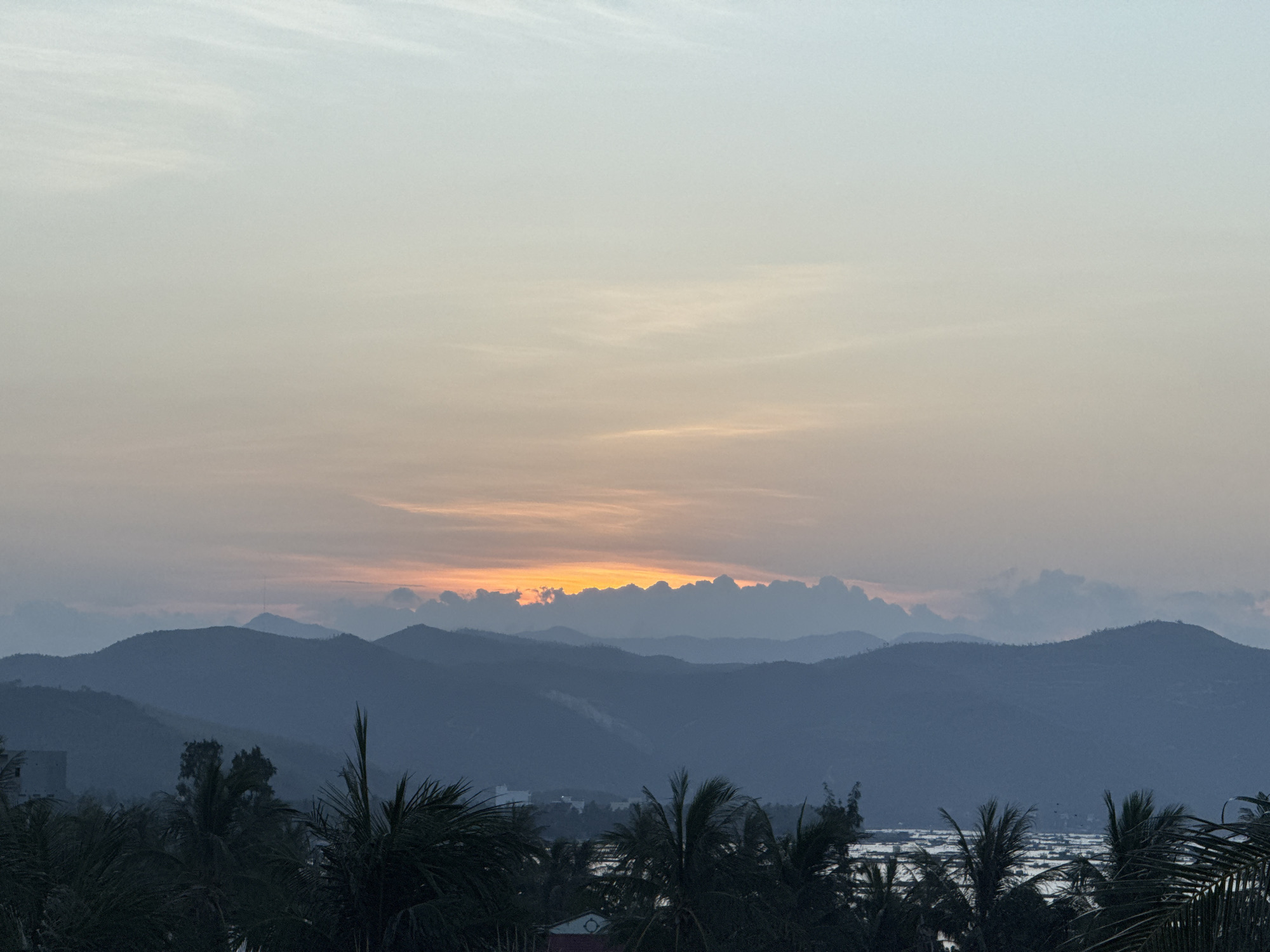 The image captures a panoramic view of the sky and mountains at sunset over the city of Song Cau, Vietnam. The sky displays a gradient of soft pastel colors, transitioning from a pale blue at the top to hues of orange and pink near the horizon. A line of silhouetted mountains forms the middle ground, with several layers of peaks receding into the distance. The foreground is dominated by the dark silhouettes of palm trees and other tropical foliage. Below the mountains, there are hints of structures and possibly water, suggesting the presence of the city and its surroundings. The overall atmosphere is serene and tranquil, characteristic of a tropical sunset.