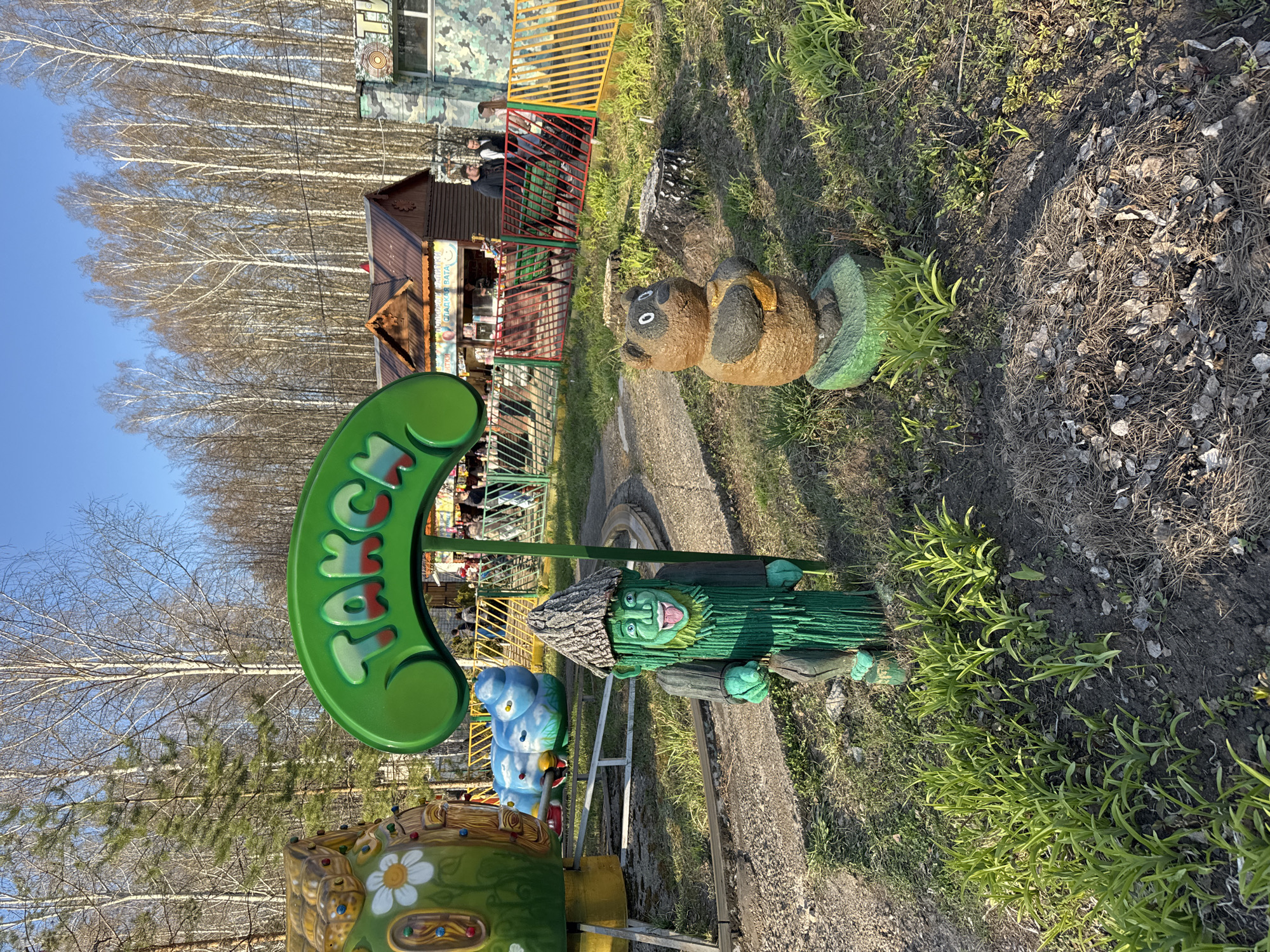 The image depicts a park scene in Chaikovsky, Russia. In the foreground, a ride is visible, featuring brightly colored characters and a green sign with Cyrillic lettering. To the right of the ride, there are decorative sculptures of a raccoon and a green goblin-like figure. The background includes a small building and a wooded area with bare trees. The sky is blue. The overall environment suggests an amusement or recreational area. There are some people standing near a fence in the background.