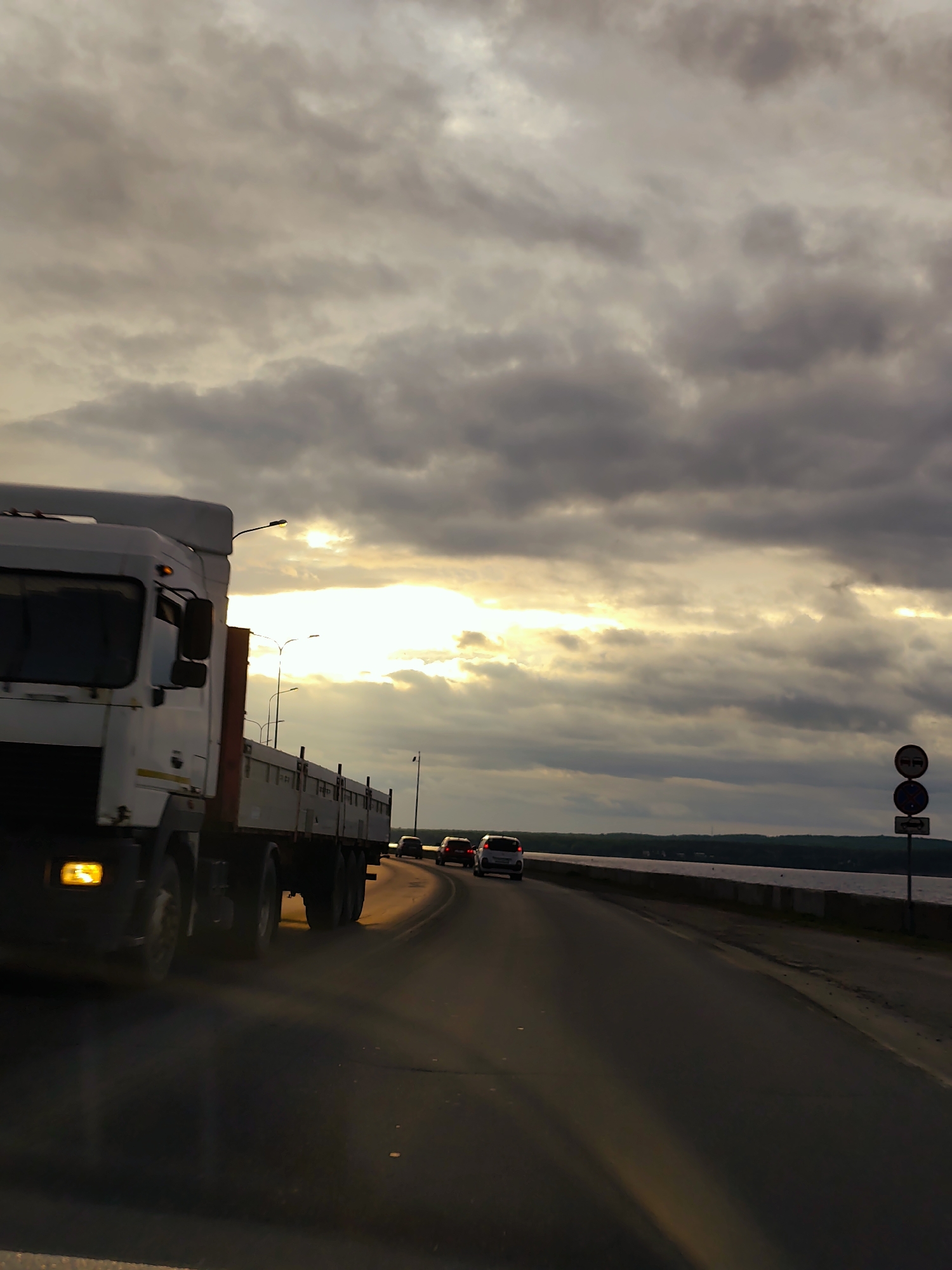 The image depicts a roadside scene in the city of Chaikovsky, Russia, at what appears to be late afternoon or sunset. A multi-lane asphalt road curves slightly to the right in the foreground and midground. On the left side of the frame, a large white flatbed truck, with some motion blur, occupies a significant portion of the foreground, traveling from left to right. Its front headlight is illuminated with a yellow glow.
Further down the road, several passenger vehicles, including a white car, are visible, also moving in the same direction. The road is bordered on its right by a low concrete barrier or wall, beyond which lies a wide body of water, likely a river or reservoir, stretching towards a distant, low-lying shoreline.
The sky is heavily overcast with numerous dark gray and brown clouds, but a prominent, bright band of golden sunlight pierces through the clouds horizontally across the mid-upper portion of the sky, casting a warm glow on parts of the scene and illuminating the underside of some clouds. Several lampposts are positioned along the road, their lights not prominently visible but some structures silhouetted against the sky. On the right side of the road, in the midground, a vertical pole displays two stacked circular road signs: the upper sign is a red circle with a white bar ("No Entry"), and the lower sign is a blue circle with a red diagonal line ("No Stopping" or "No Parking"). No individuals are explicitly identifiable in the scene.