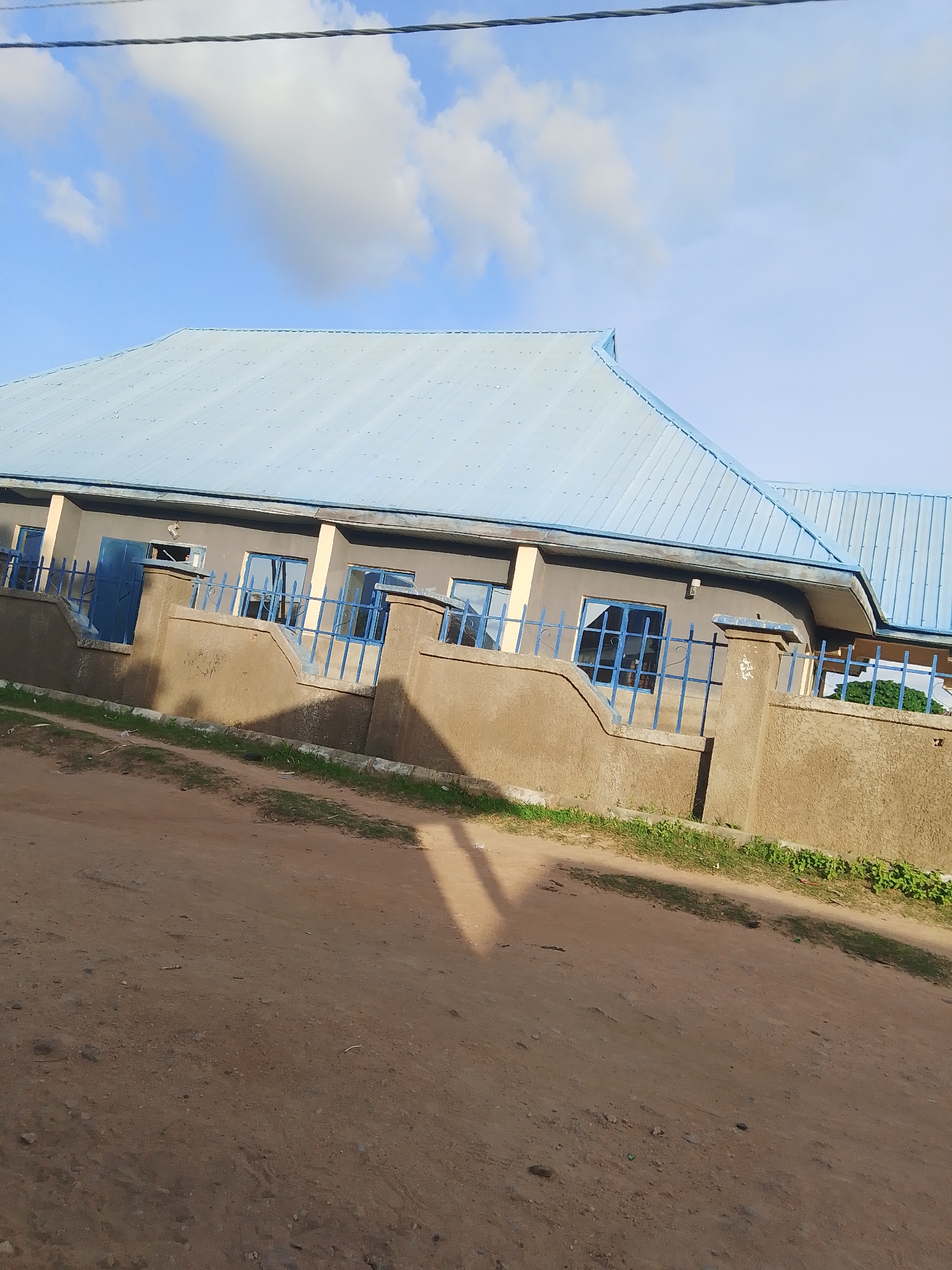 In Jalingo, Nigeria, a building with a light blue, corrugated metal roof stands under a partly cloudy sky. The building has a gray facade with beige decorative elements around the windows and at the roofline. Several blue-framed windows and a blue metal gate are visible. The building is surrounded by a low, light brown concrete wall topped with a blue metal fence. The ground in front of the building is a dirt road with patches of green grass and some scattered debris. A shadow from a power line is cast across the scene.