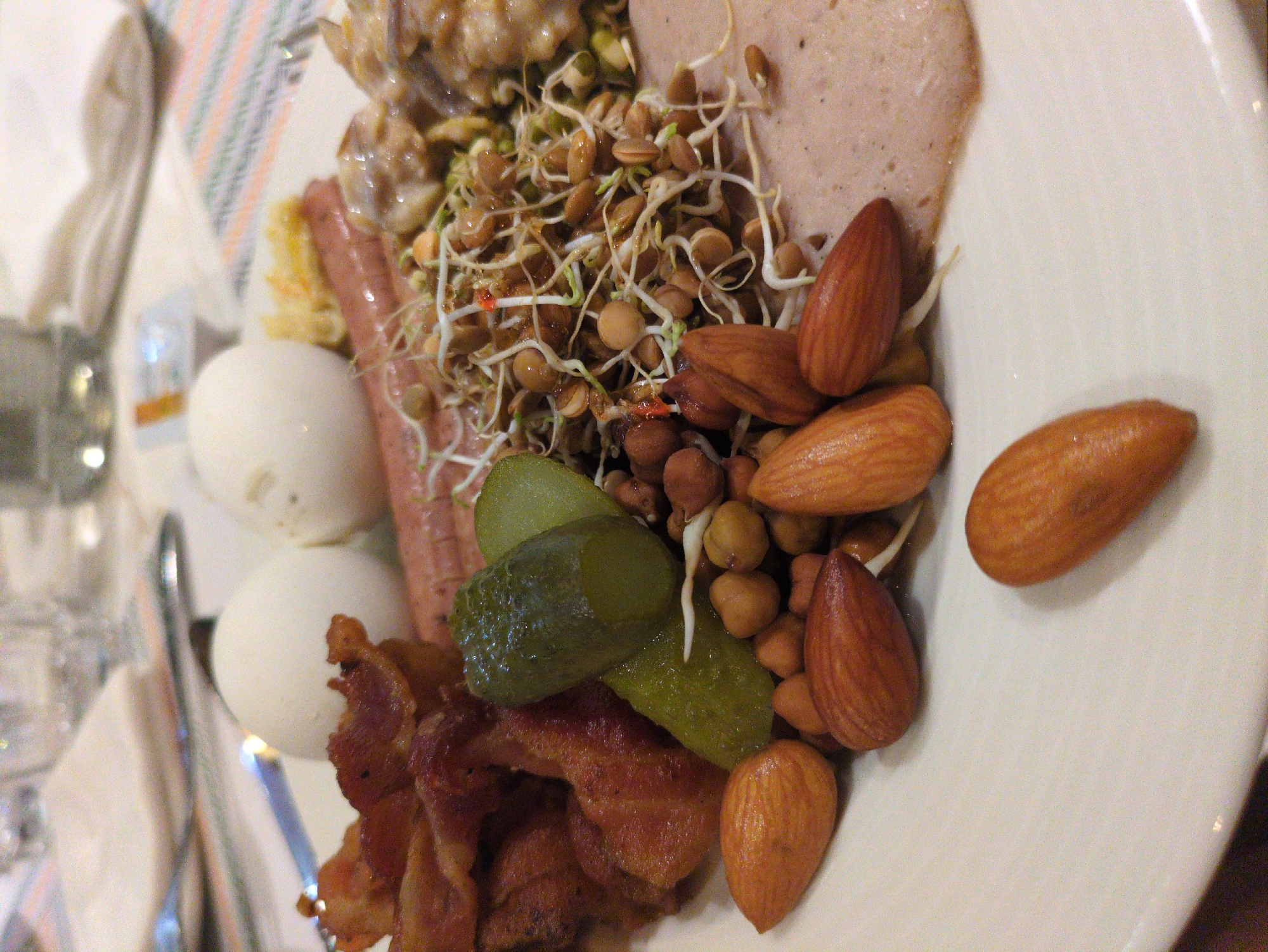 An overhead medium shot captures a white plate filled with a variety of food items, likely part of a breakfast or buffet spread. The food is arranged in distinct sections. On the left side of the plate, several strips of crispy, fried bacon are piled high. Adjacent to the bacon are two pickle spears, one whole and one sliced, with a glistening, green surface. Below and interspersed with the pickles are small, brown beans and sprouts, creating a textured mound. Further to the right, two whole, white-shelled eggs are visible. In the background, a partially visible pink sausage lies horizontally, and to its right, a portion of what appears to be sliced ham or other processed meat is present. There is also a small scattering of what looks like shredded cheese or another yellow food item near the sausage. The overall impression is a diverse culinary offering. The image does not provide any information about the location in Agra, India.