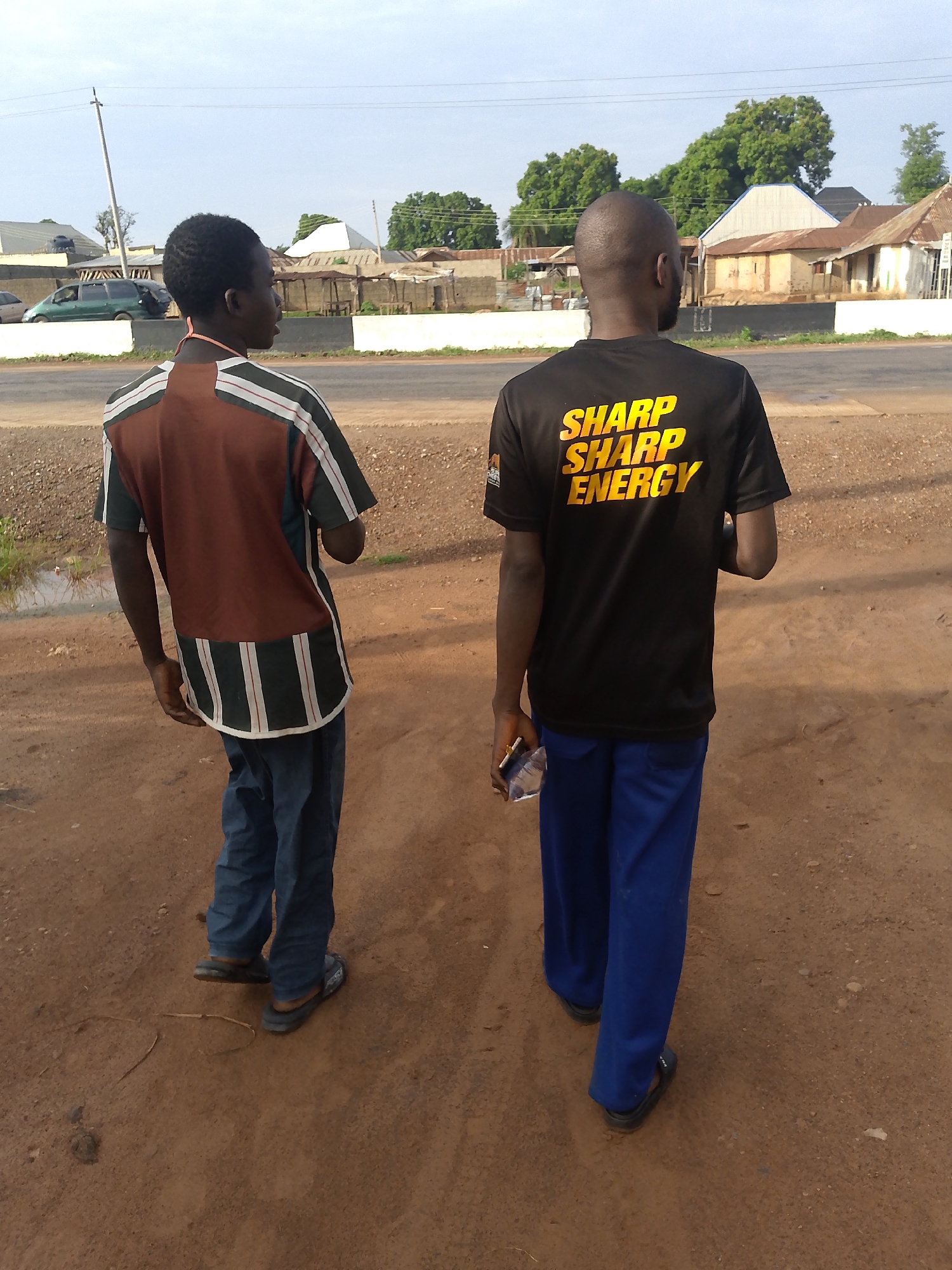 Two young men are standing on a dirt road in front of a street in Keffi, Nigeria. The man on the left is wearing a brown and white striped shirt and blue pants. The man on the right is wearing a black t-shirt with yellow lettering that reads "SHARP SHARP ENERGY" and blue pants. Both men are facing away from the camera. In the background, there are several buildings and trees. The sky is cloudy.