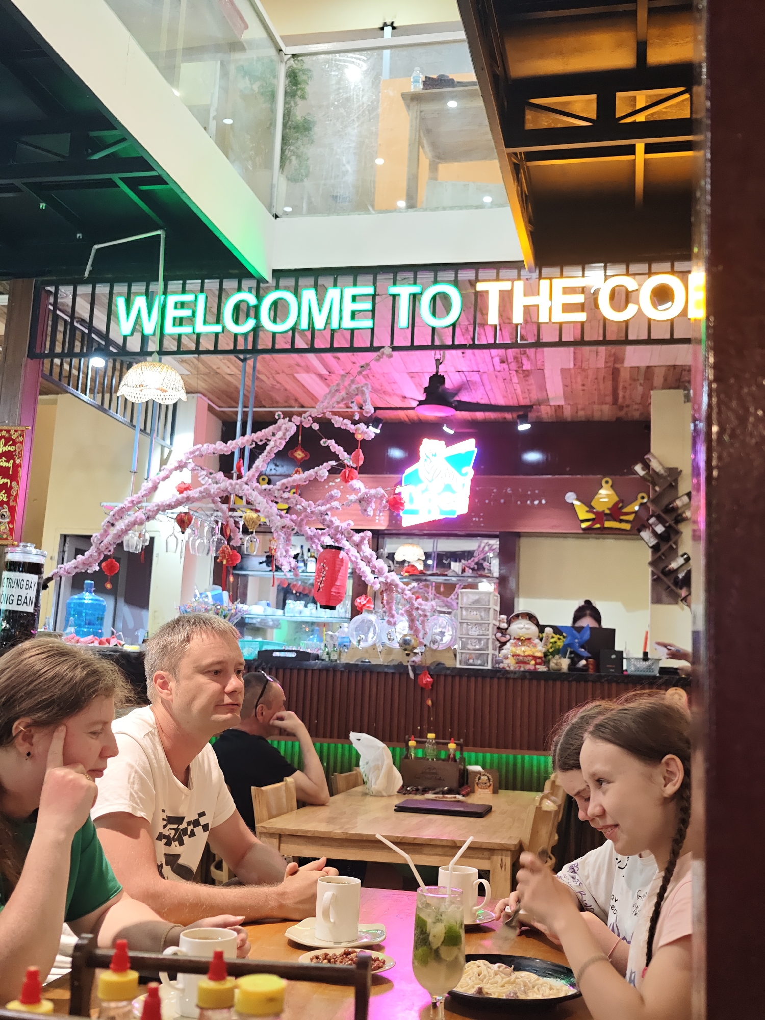 The image depicts a group of people seated at tables inside a restaurant. The setting appears to be a casual dining establishment, possibly a cafe, given the "WELCOME TO THE COF" neon sign visible overhead. The decor includes decorative pink cherry blossom branches with hanging ornaments and lanterns, suggesting a festive or seasonal theme.
In the foreground, three individuals are visible. On the left, a woman with brown hair in a green shirt looks thoughtfully away from the camera. Beside her, a man with fair skin and short hair, wearing a white t-shirt, is also looking away. To the right, two young girls are seated, one with her hair in braids, looking towards the table. One of the girls is holding a utensil and appears to be interacting with food on her plate.
On the table in front of them, there are drinks, including a tall glass with a green beverage and ice, and mugs of coffee. There is also a plate of pasta and some small bowls containing what appear to be nuts or snacks. In the background, other patrons and staff are visible, as well as bar area with bottles and decorative items, including a golden crown emblem. A water cooler and a jar with Vietnamese text are also present in the midground.
The lighting suggests it might be evening or indoors with artificial lighting. The presence of the neon sign and the indoor setting indicate it is not an outdoor scene. The overall atmosphere appears relaxed and social. Visible text includes "WELCOME TO THE COF" on the neon sign and Vietnamese text on a jar in the midground, partially obscured.