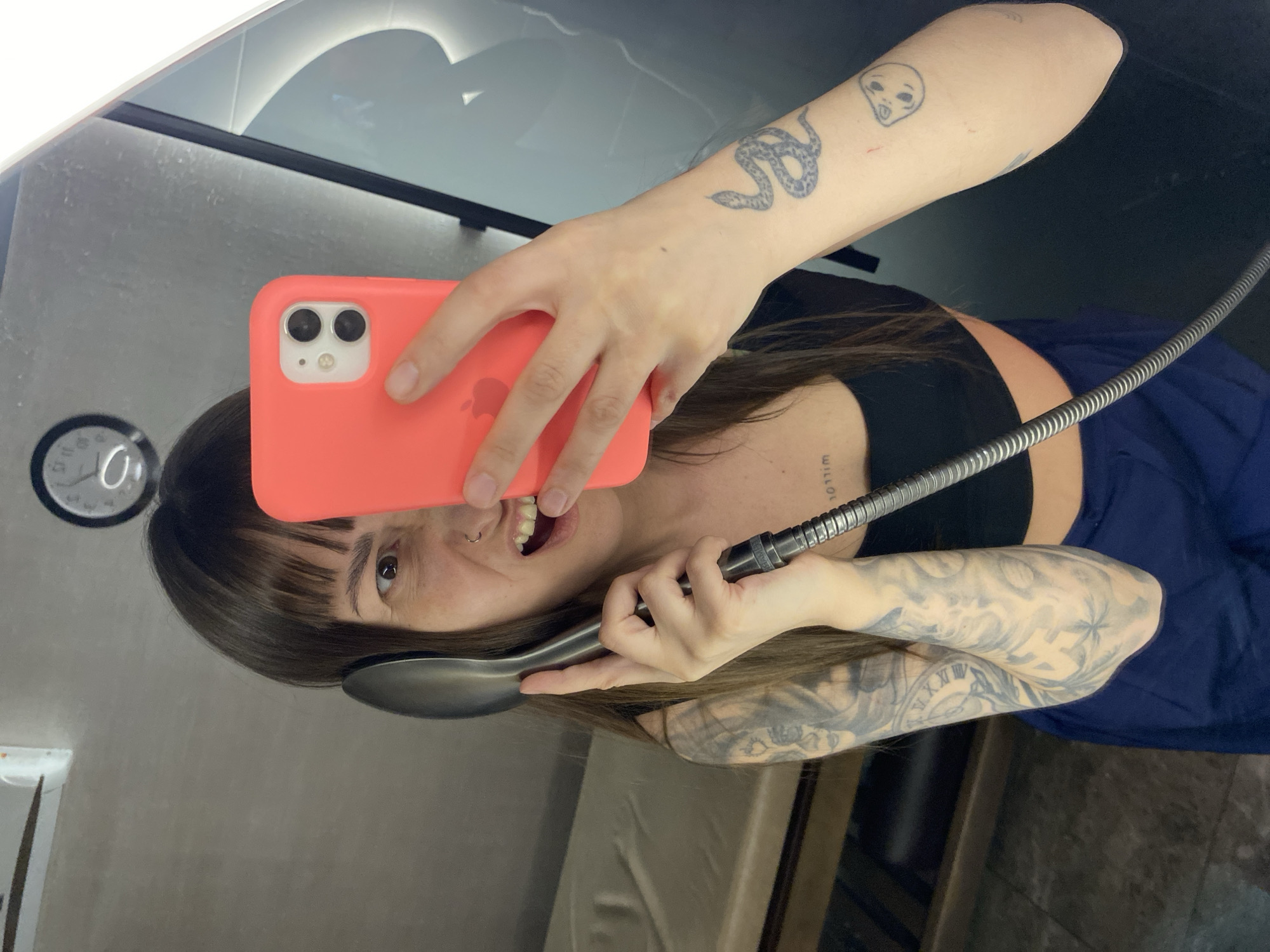 A playful mirror-selfie captures a person with dark hair and bangs, holding a coral-colored iPhone in their right hand. They are taking the photo while holding a large, shiny metal spoon to their left ear as if it were a telephone receiver. A flexible metallic hose or cable is also held in their left hand, near the spoon. The person has a wide, open-mouthed smile, revealing teeth, and a nose ring.
They wear a black crop top and a blue skirt or loose pants. Their arms are extensively tattooed: a small snake on the right forearm, an alien head on the right bicep, and a full, intricate sleeve tattoo on the left arm featuring patterns that include Roman numerals (e.g., V, X, XI, XII). A small text tattoo reading "will..." is visible on their neck/collarbone area.
The setting appears to be indoors, reflected in what seems to be a mirror. The background shows a modern-looking room with light surfaces and a distinct curved light fixture or design element overhead. To the left, a circular object resembling a clock or gauge with markings like "0," "3," "6," and "9" is partially visible. The overall mood of the image is cheerful and lighthearted.
Location context: Votkinsk, Russia.