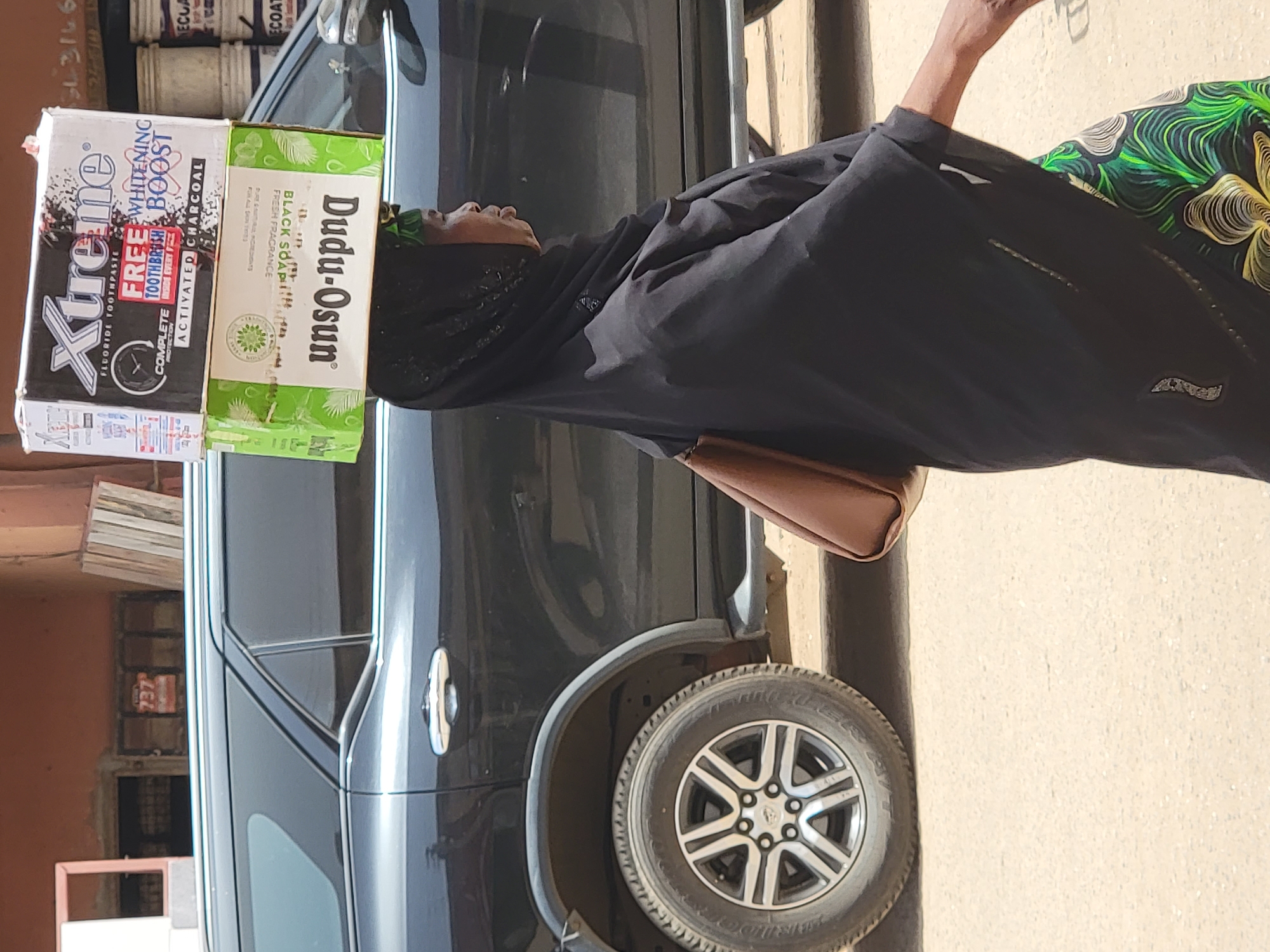 A woman in Maiburiji, Nigeria, wearing a black hijab and a patterned green skirt, is standing next to a black SUV. Two boxes are balanced on her head. The first, on the left, is a white and black box of Xtreme brand fluoride toothpaste, with "WHITENING BOOST" and "ACTIVATED CHARCOAL" written on it. The second box, on the right, is green and labeled "Dudu-Osun BLACK SOAP" with "FRESH FRAGRANCE." A brown purse hangs from her shoulder. A stack of books is visible behind the SUV, along with paint cans stacked on a shelf. A red sign with "737" is on the wall behind the SUV. The ground is light brown.