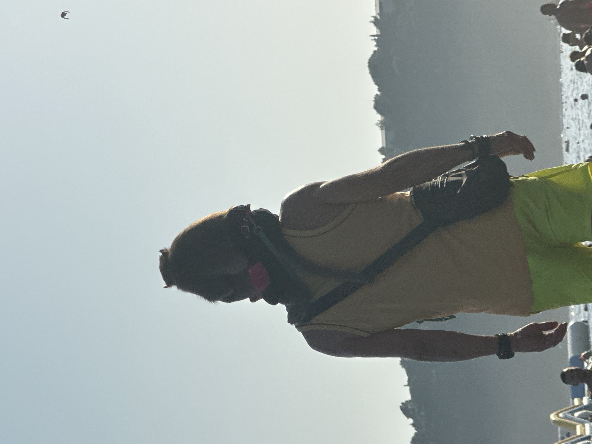 The image shows a person standing on a beach in Boreti, Montenegro. The person is viewed from behind, wearing a yellow tank top and neon green shorts, with a black sling bag across their body. The person has dark hair and is wearing a black face mask. The background is a hazy landscape of hills with buildings on them, and the sea with people swimming. The sky is clear and bright.