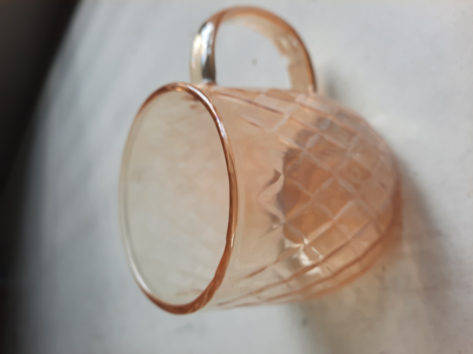 A translucent, pale orange glass mug with a diamond-patterned texture and a looped handle is positioned on a textured, light-colored surface. The background is out of focus, showing a gradient from dark gray to light gray. There is no indication of the city of Lhokseumawe, Indonesia, in this image.