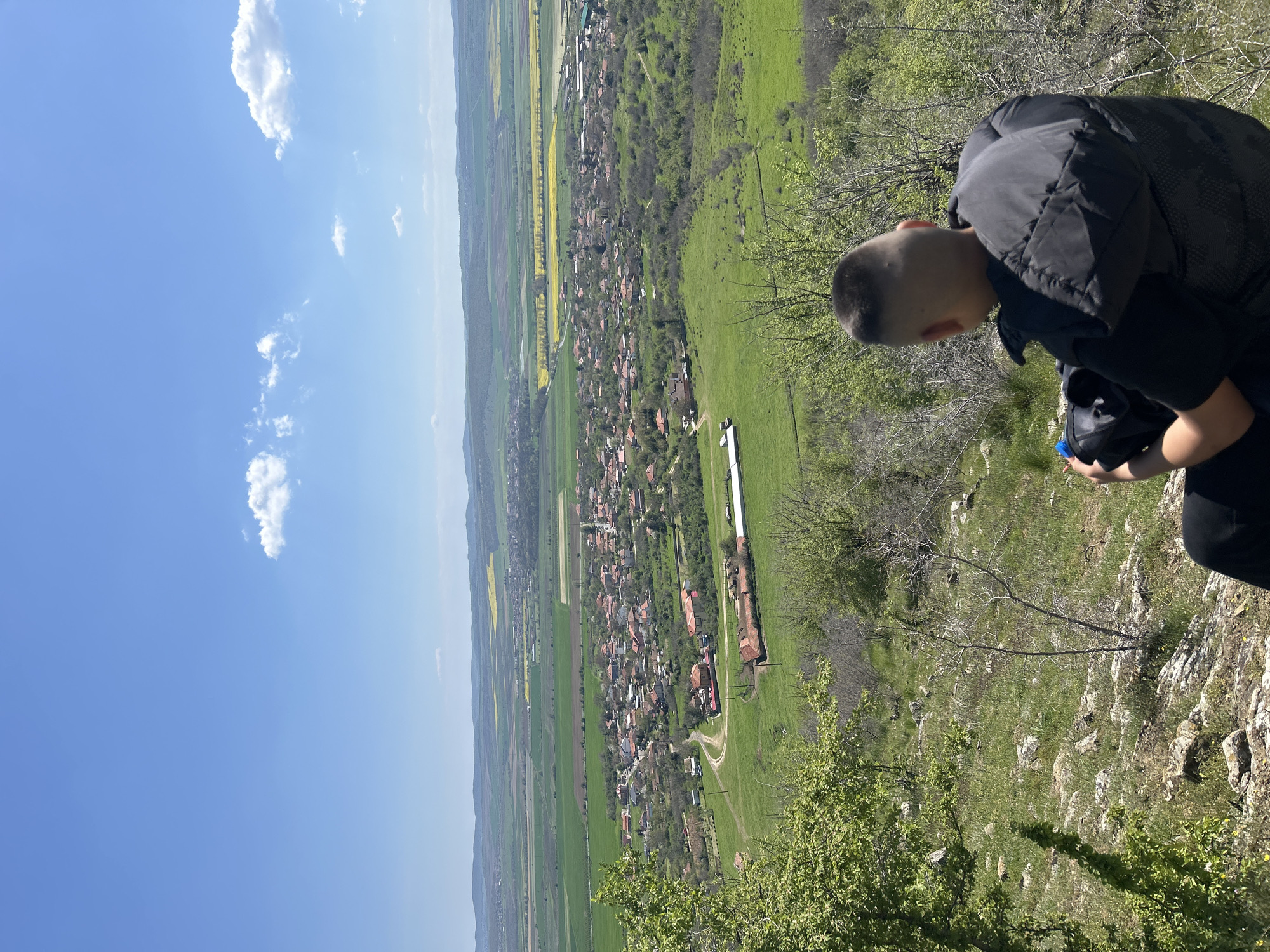 A person stands on a rocky slope overlooking a village nestled within a landscape of green fields and scattered trees. The sky is a clear blue with a few scattered white clouds. The village has red-roofed houses and a few longer buildings near the center. In the distance are fields of bright yellow flowers interspersed with green fields. The person is wearing a dark jacket. The overall setting suggests the location is Goritsa, Bulgaria, where a village is surrounded by a landscape of fields, trees, and rocky terrain.