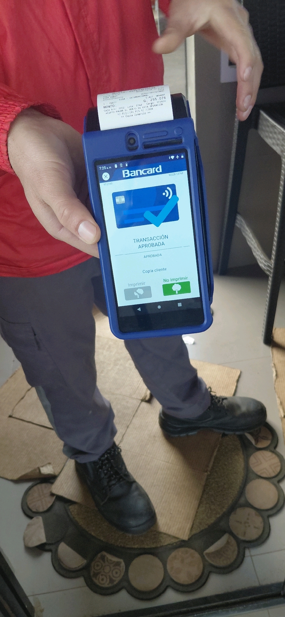 An individual wearing a red long-sleeved shirt, gray pants, and black work shoes stands on a patterned circular mat. The individual is holding a blue device with a touchscreen and a receipt printer. The screen displays "Bancard" and a message indicating "TRANSACCION APROBADA" (Transaction Approved) with a green checkmark. The receipt shows details of a credit card transaction, including "VISA DEBIT", "INTERNACIONAL", and "MONTO: 255.076". The scene appears to be indoors, with a tiled floor visible. The location is identified as Encarnacion, Paraguay.