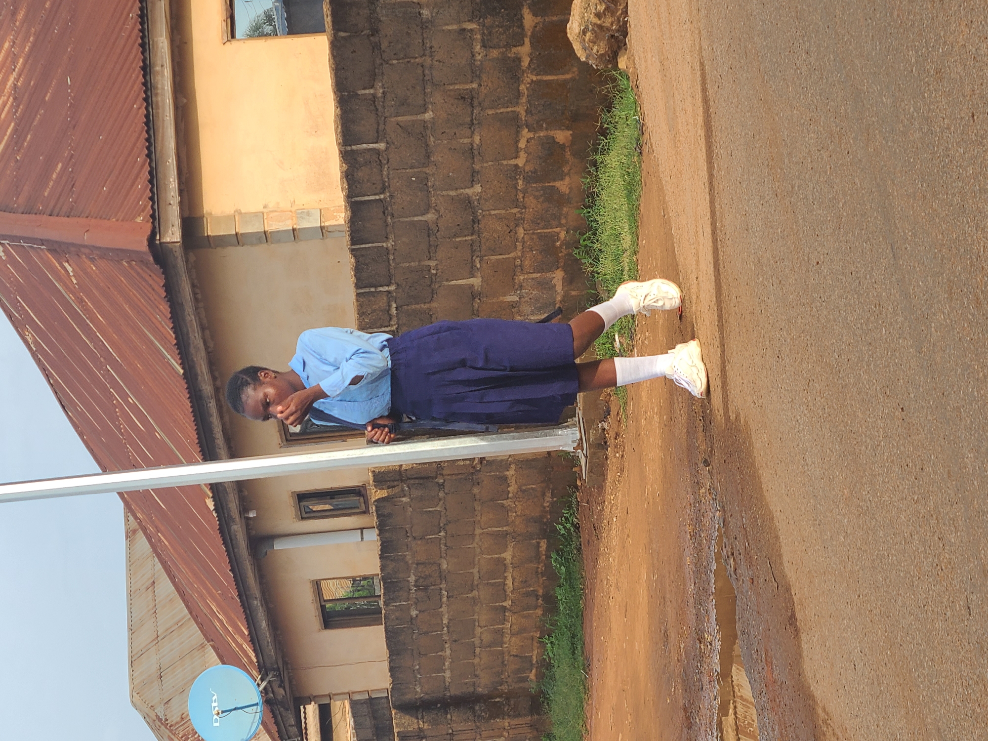A young person in a blue school uniform, white socks, and white shoes stands on a dirt and paved road next to a brick and tan building with a rusty corrugated metal roof. The person is holding their hand to their face. A utility pole with a blue DSTV satellite dish is visible near the building. The scene is located in Jos, Nigeria.