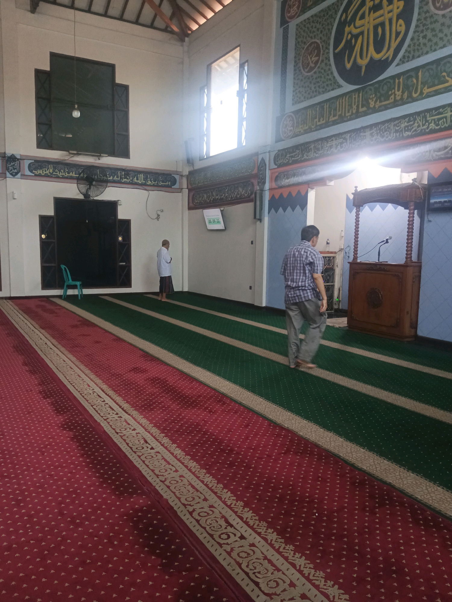 The image shows the interior of a mosque in South Tangerang, Indonesia. The mosque features red and green carpets with decorative borders. In the foreground, a man wearing a plaid shirt and grey pants walks away from the camera. In the middle ground, a man in a white shirt and sarong stands facing a wall. A wooden podium with a microphone is visible on the right side of the image. The walls are decorated with Arabic calligraphy. A digital clock displaying "00:09:32" is mounted on the wall. The ceiling is made of wood. The lighting suggests it is daytime, with natural light entering through windows.